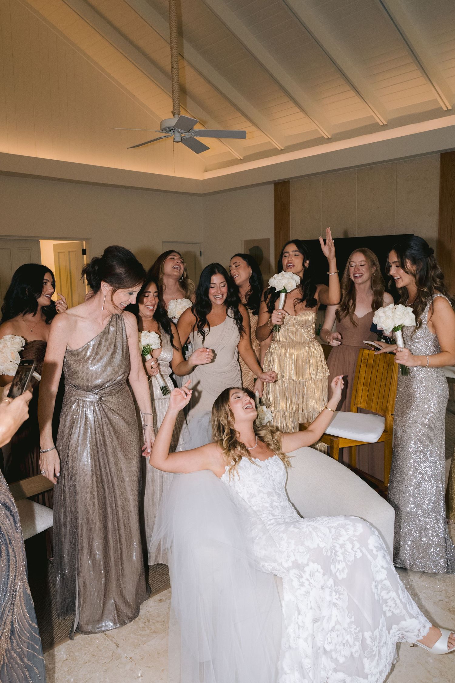 Wedding party wearing elegant gowns in shades of silver and champagne dance together at reception celebration.