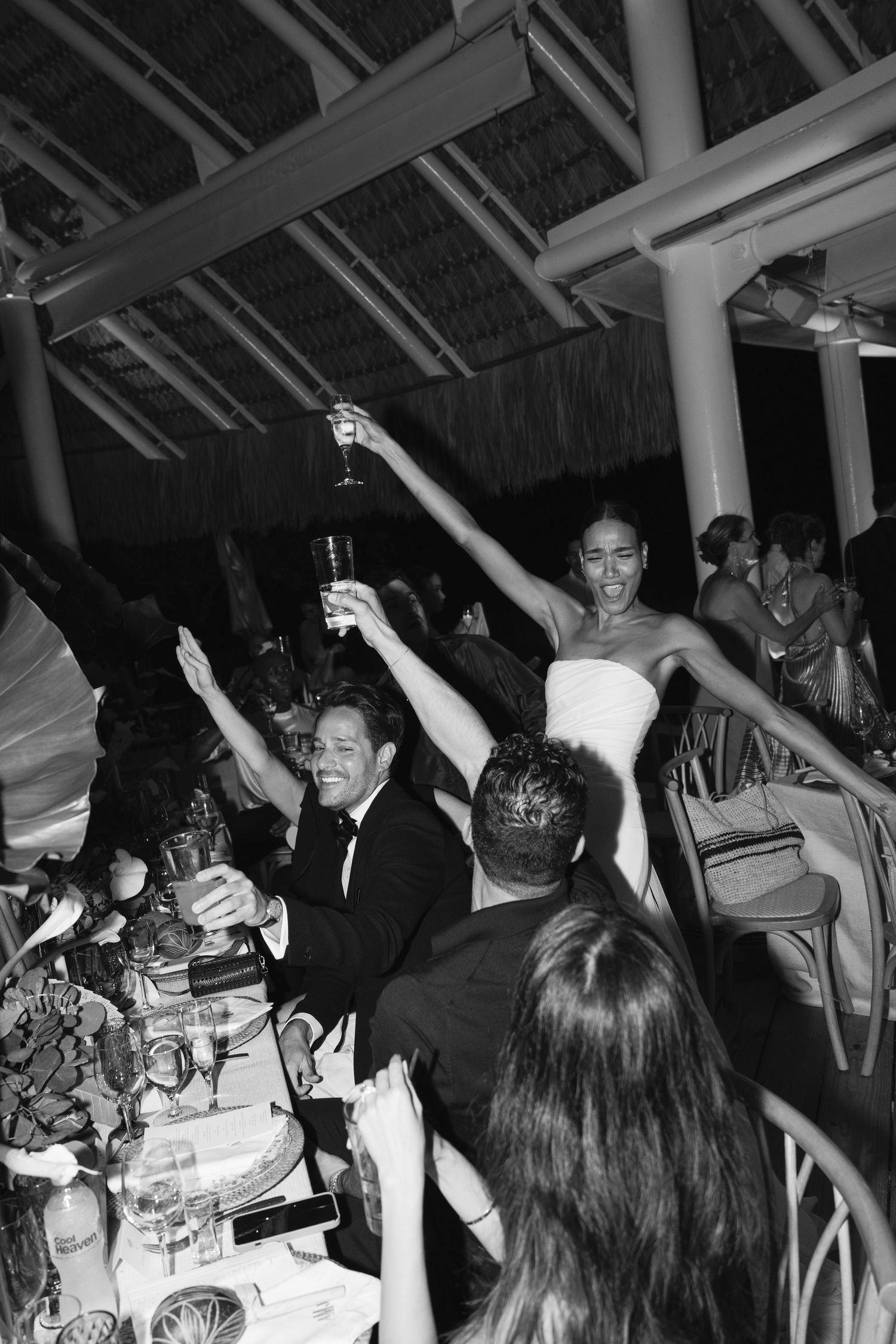 People celebrating at a lively dinner party with raised glasses and drinks in an indoor venue at night.