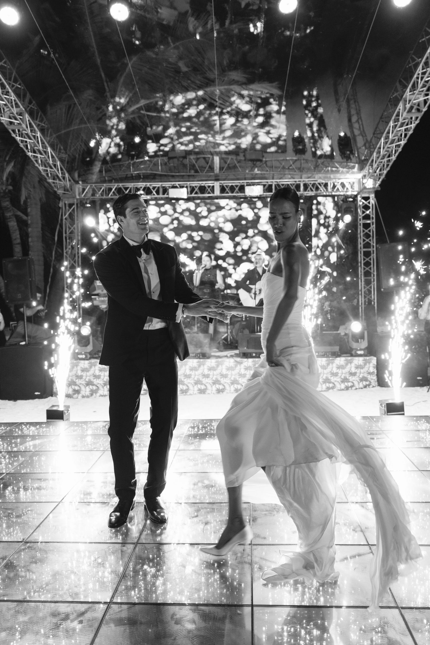 A bride and groom dancing together on a lit dance floor with sparkling lights and stage rigging in the background.