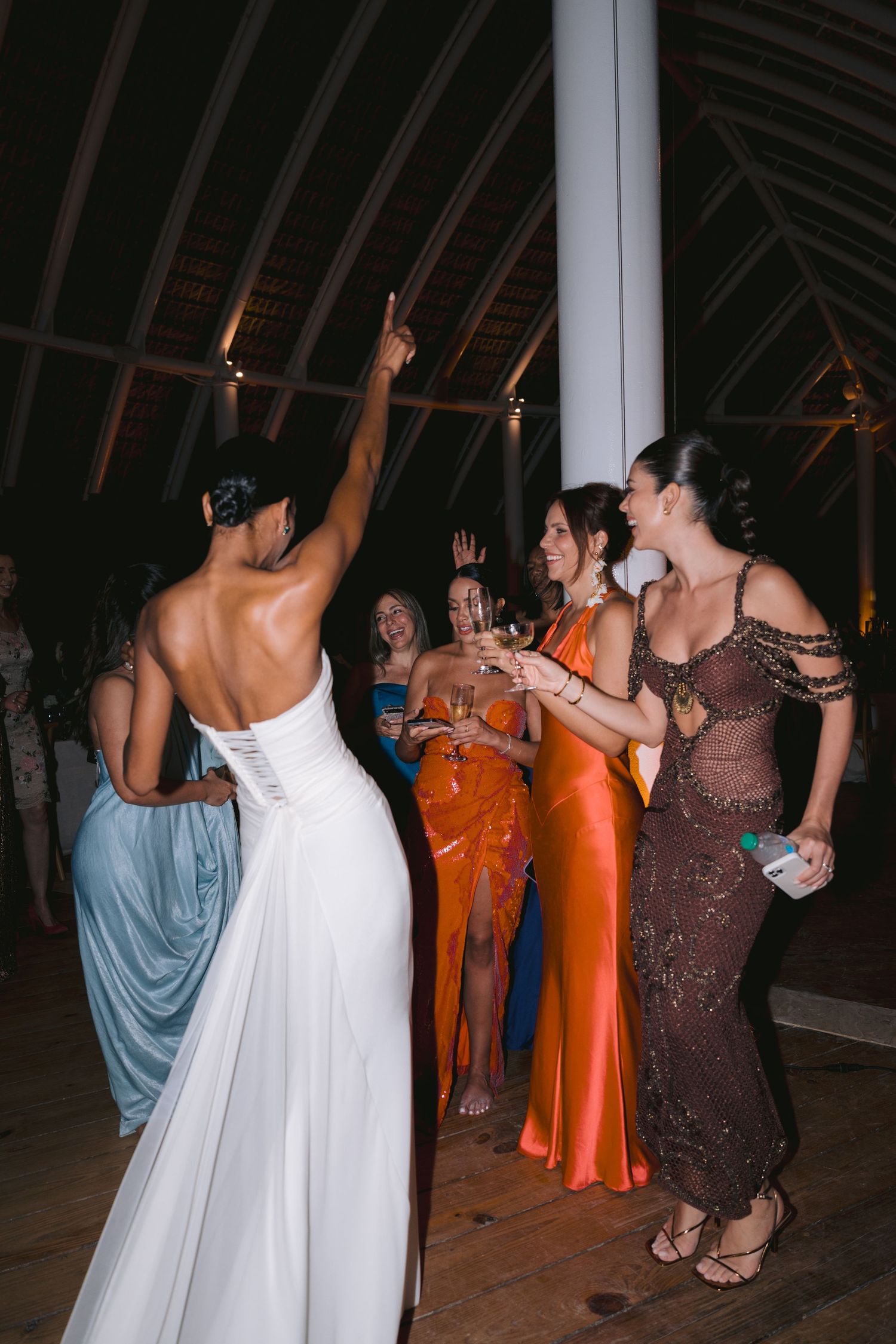Party guests in formal evening wear dance and celebrate on a wooden floor under high ceilings at night.