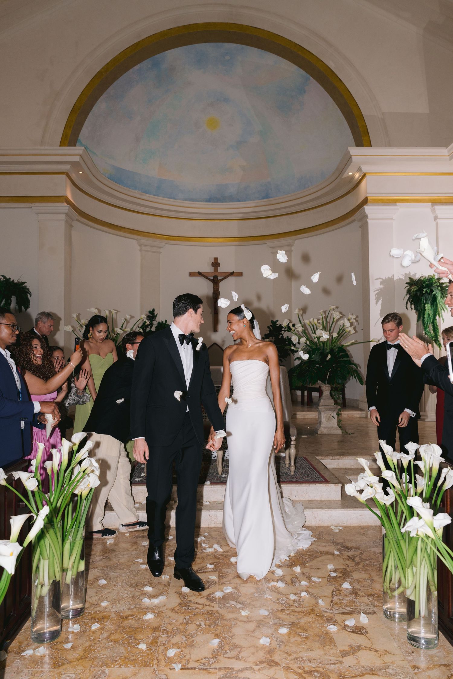 Newlyweds walk down church aisle decorated with white lilies as guests celebrate with confetti and petals.