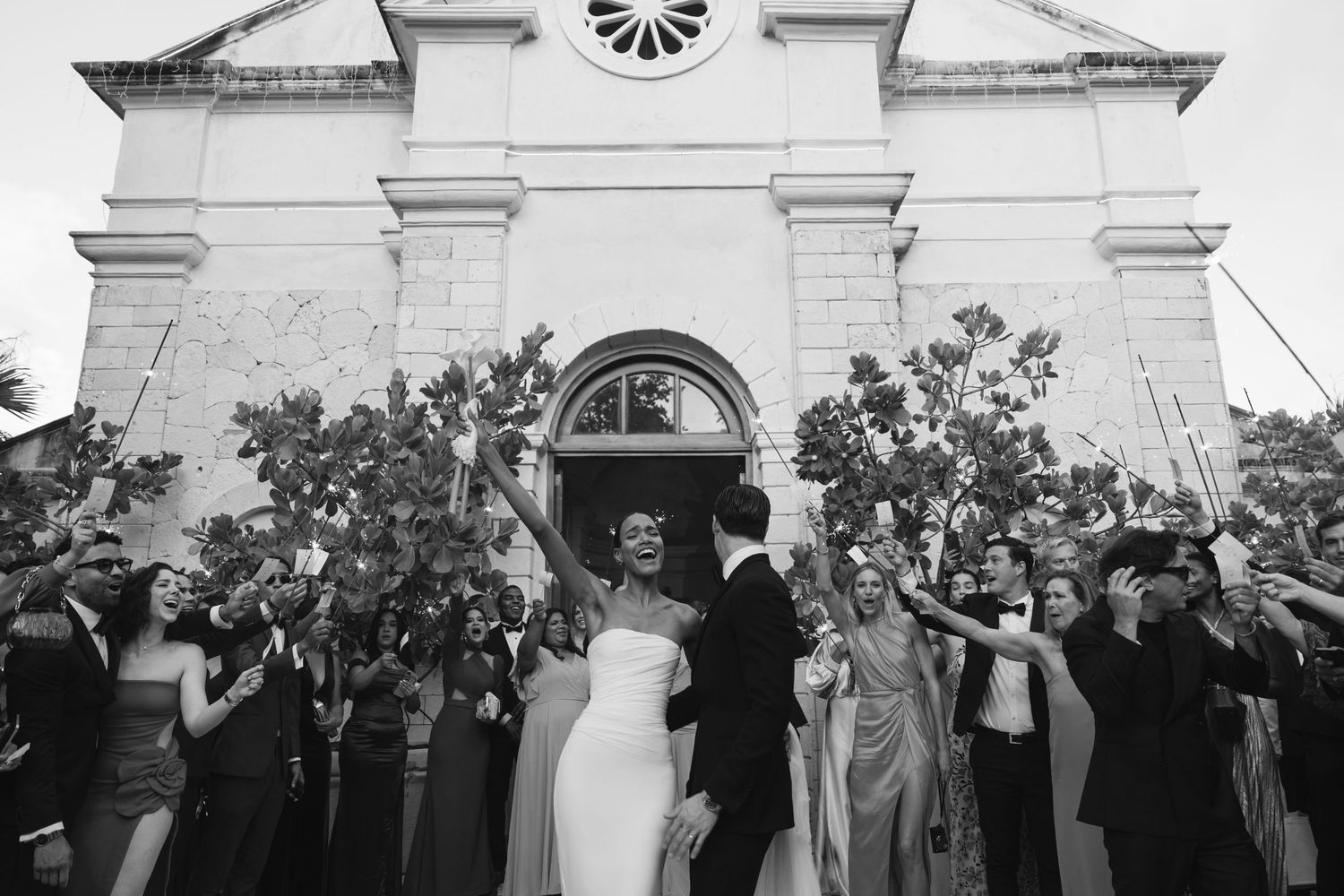 Wedding guests celebrate with rose petal toss as newlyweds exit historic church with arched doorway in black and white photo.