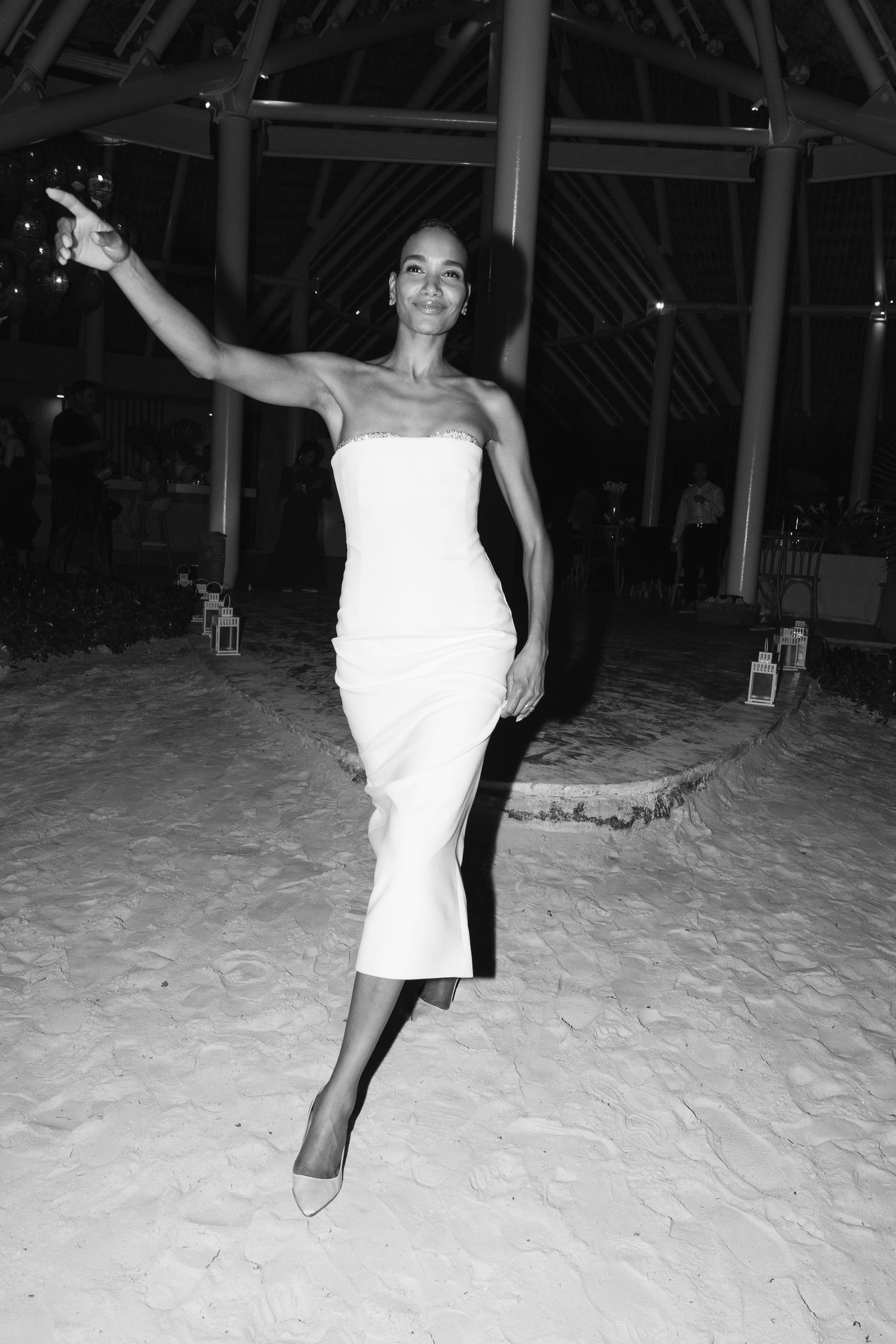 A graceful figure in a white strapless dress dances on sandy ground beneath a thatched roof structure at night.