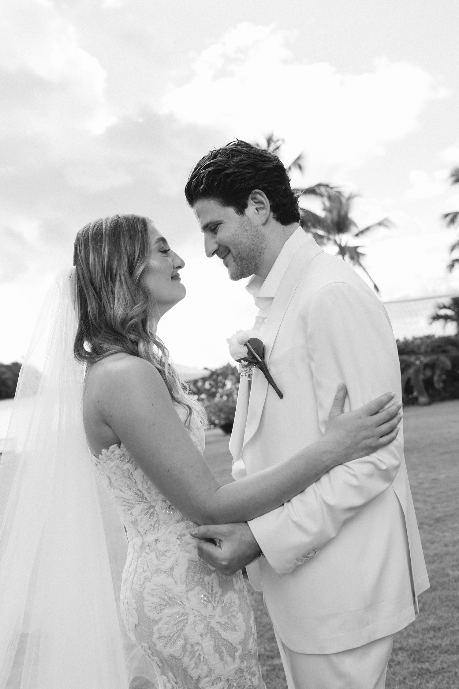 A romantic black and white wedding photo of a couple embracing outdoors with palm trees and clouds in the background.
