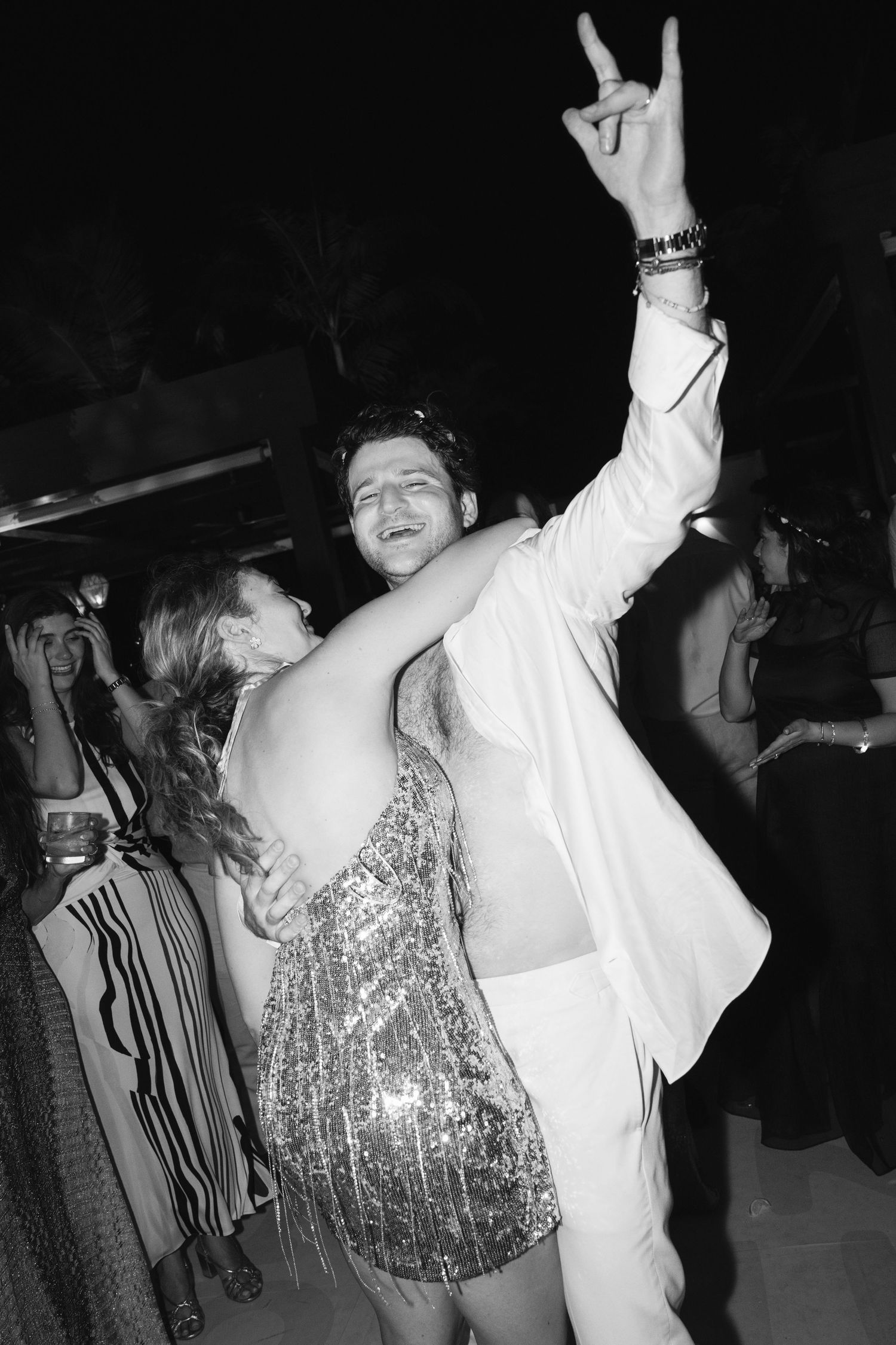A couple dances energetically at a party, with one person in a sparkly dress and another in a white suit.