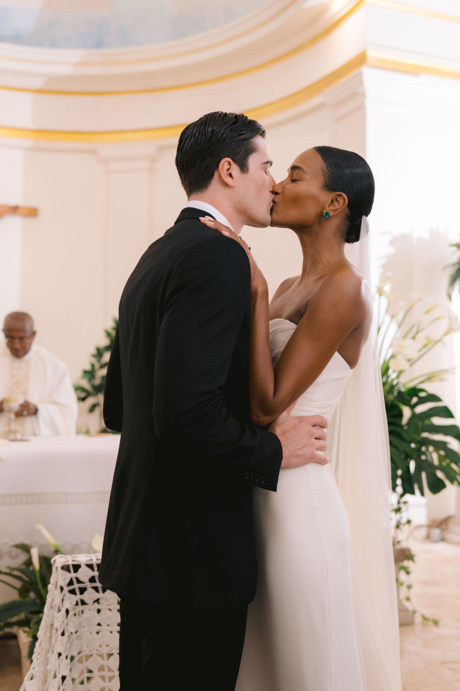 A romantic wedding kiss between a couple in a white room decorated with tropical monstera leaves and elegant decor.