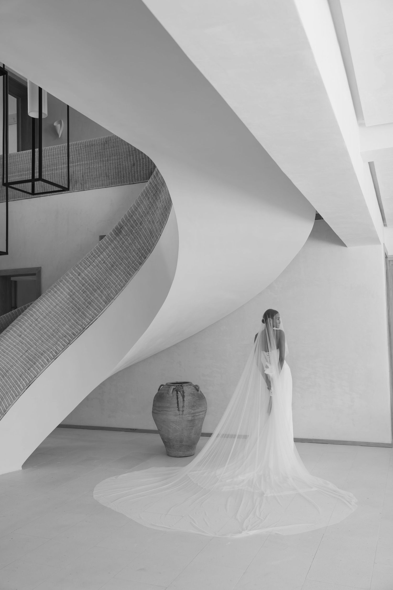 Elegant bridal figure in white dress with flowing cathedral veil stands beside curved staircase and large pottery vase.