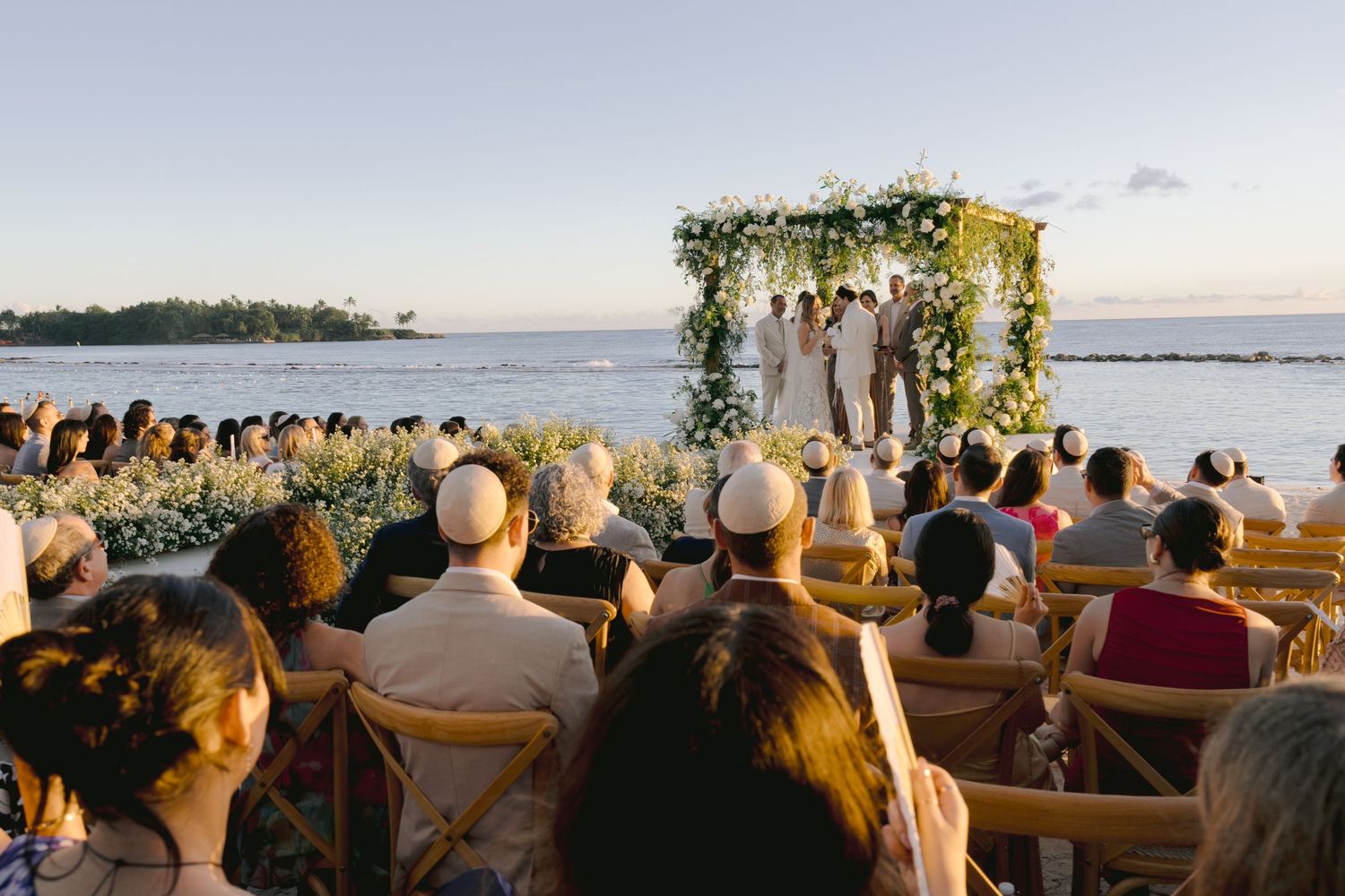 Wedding ceremony guests seated outdoors watching a beachside celebration at sunset.
