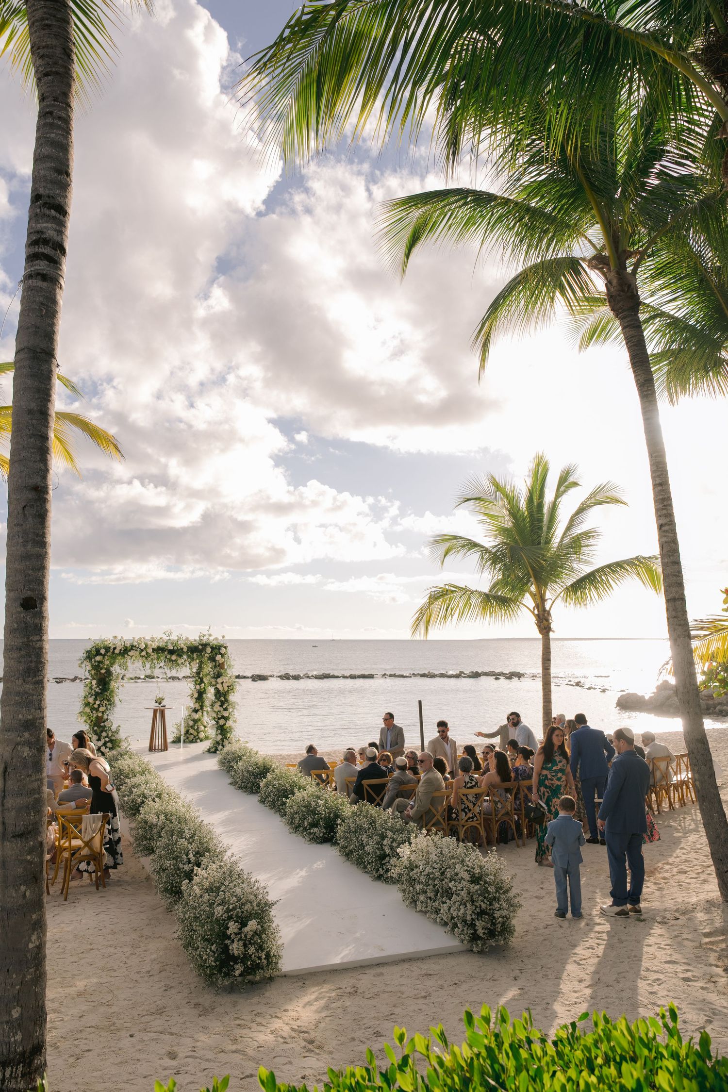 Tropical beach wedding ceremony setup with palm trees and white chairs arranged on sandy shore at sunset.