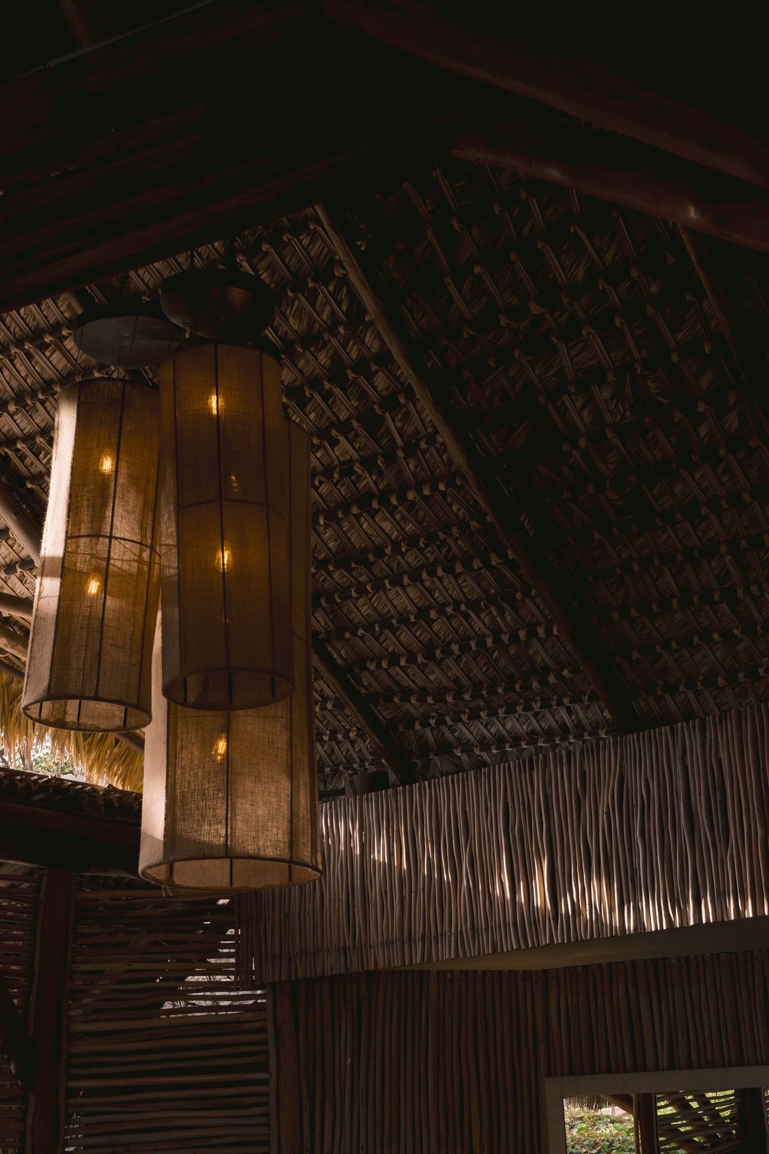 Wooden lantern hangs from bamboo and thatch ceiling in tropical style restaurant interior with moody lighting.