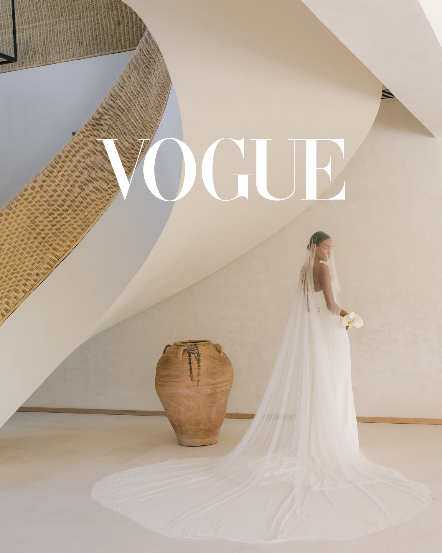 A bride in a flowing white gown and veil stands in an elegant minimalist space with curved walls and a large ceramic vase.