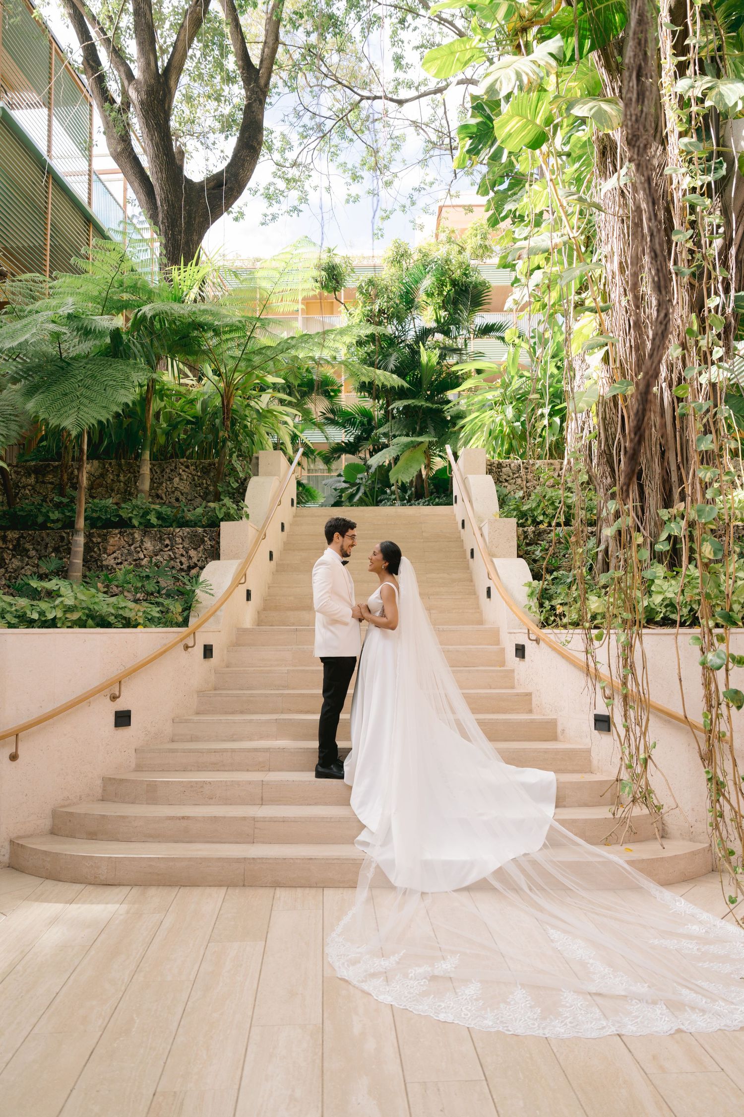 A wedding couple stands on elegant white stairs surrounded by lush tropical gardens.