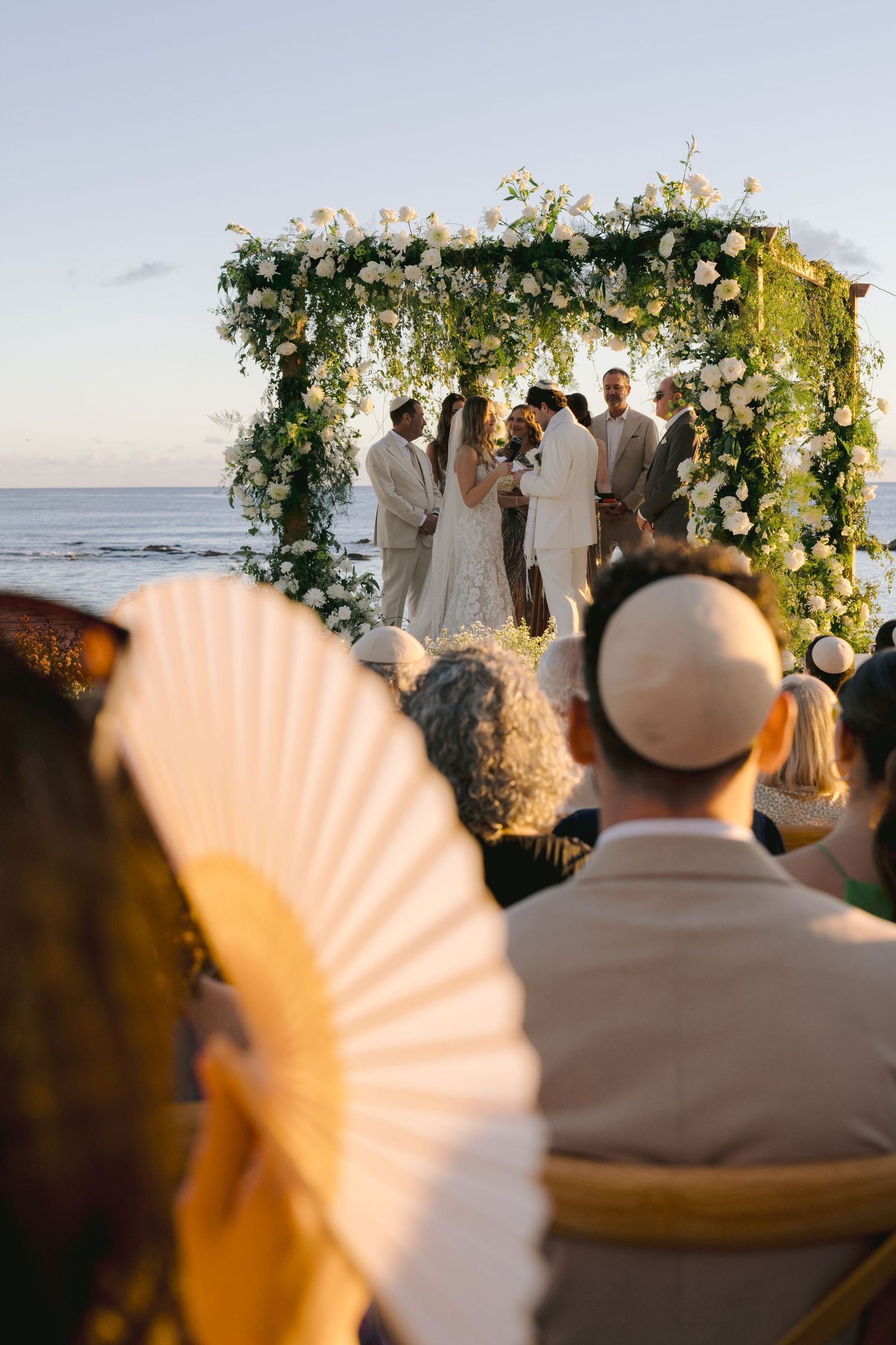 A beachside wedding ceremony with floral arch decorations and guests seated with paper fans at sunset.