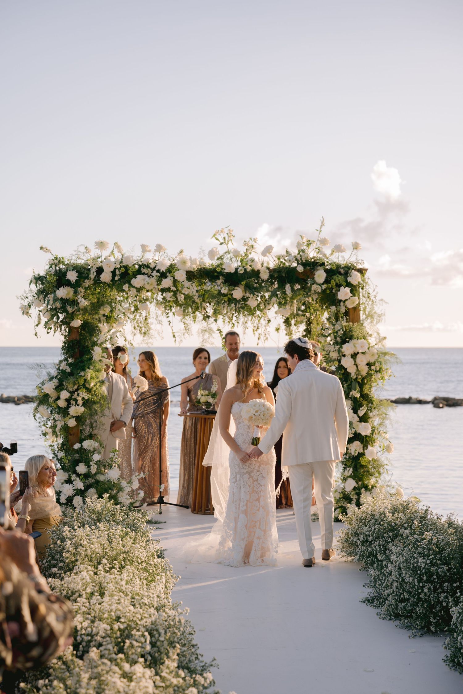 Elegant beachfront wedding ceremony setup with white floral arch and guests gathering at sunset by the ocean.