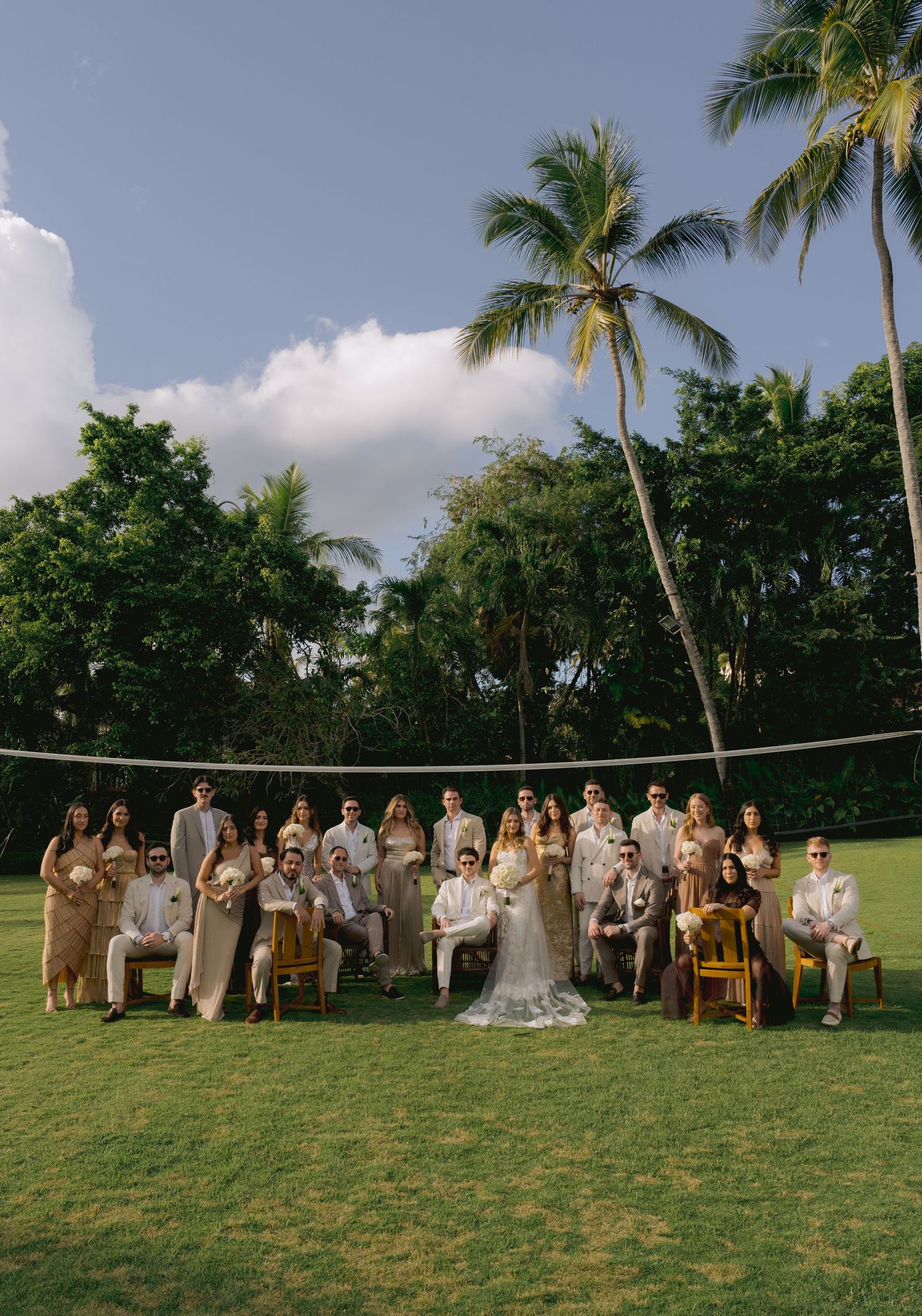 Wedding party members gather on a lush green lawn with palm trees for outdoor group photos in tropical setting.