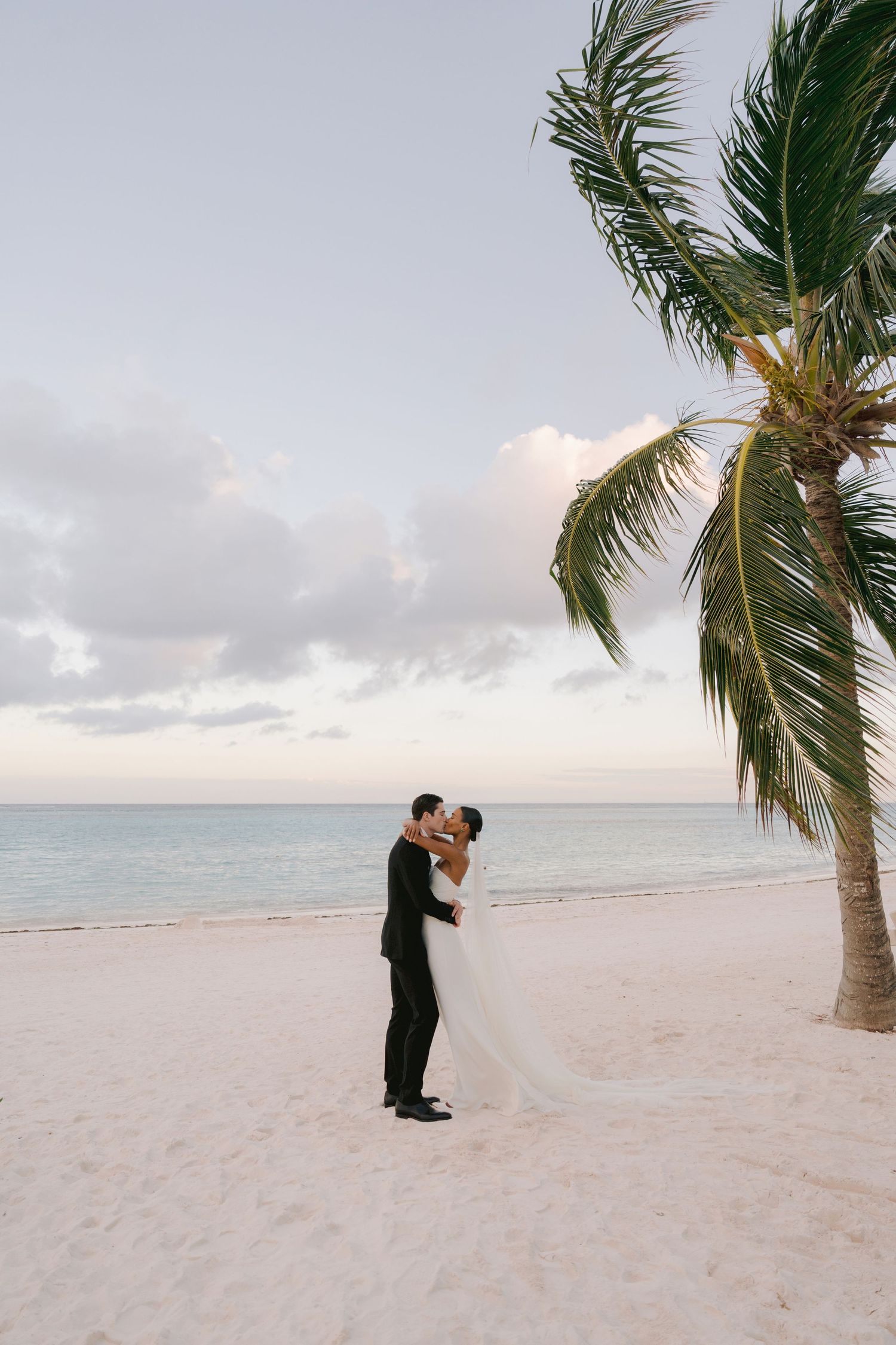 A romantic beach wedding scene with a couple embracing under a palm tree during sunset on a white sand shoreline.