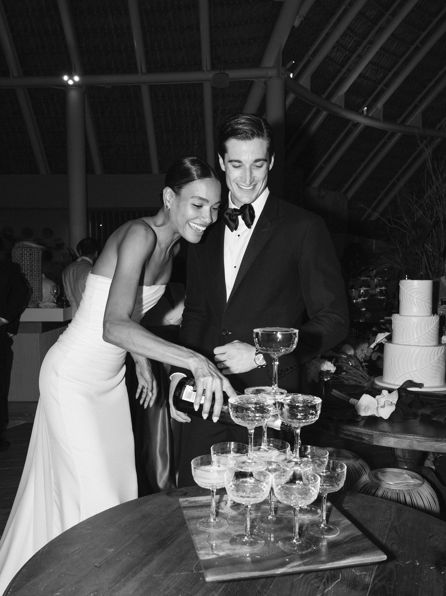 A couple in formal attire pours champagne into a tower of glasses at an elegant wedding reception.