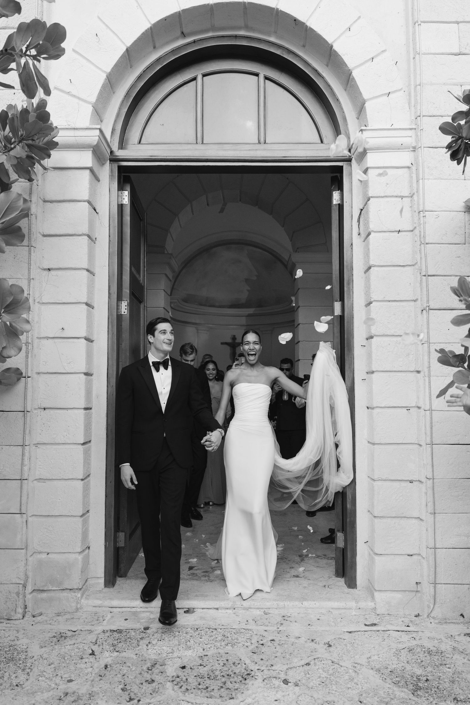 A couple exits through an elegant arched church doorway in formal wedding attire while guests celebrate behind them.