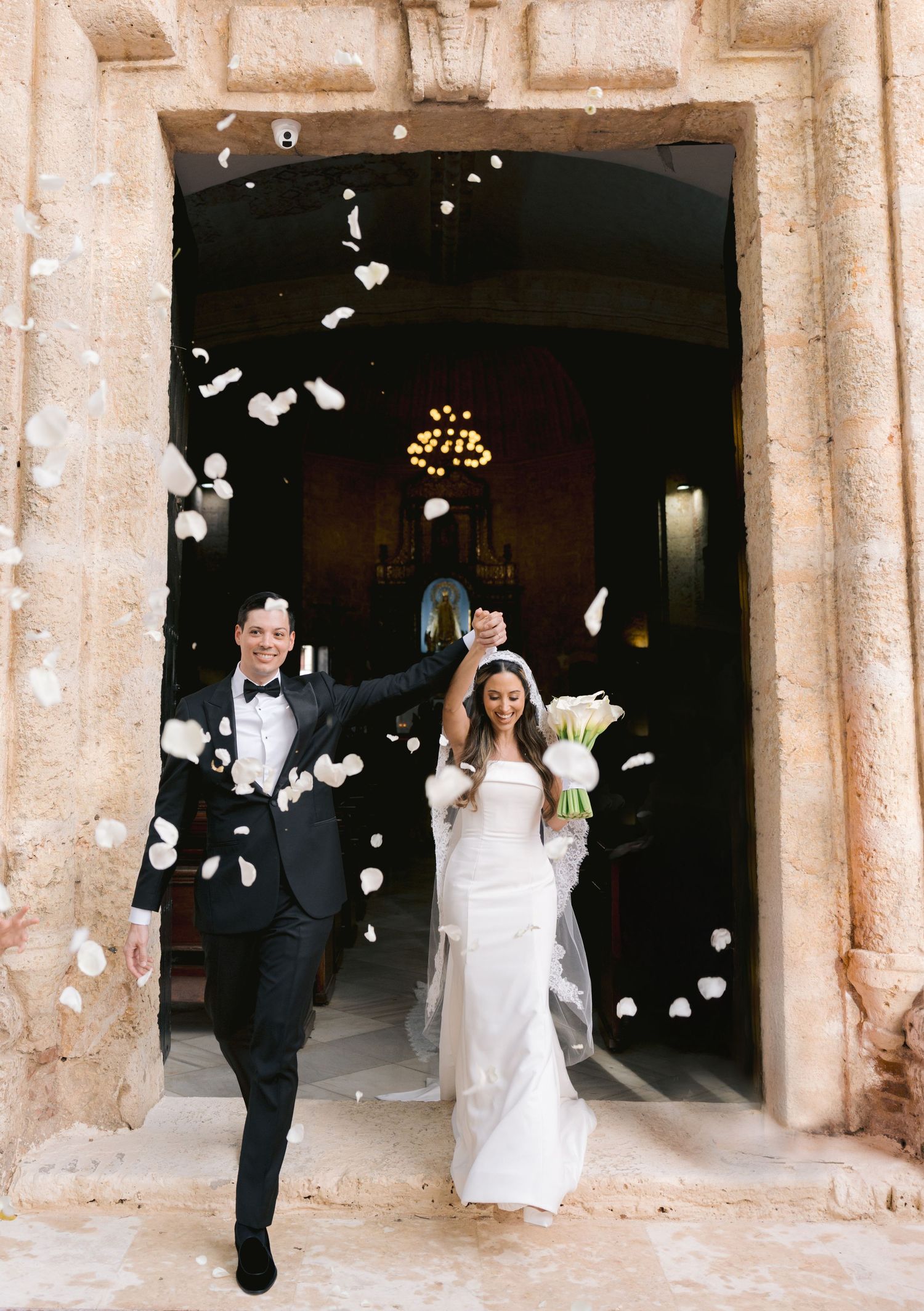 A wedding couple exits a church while white flower petals are being thrown in celebration.