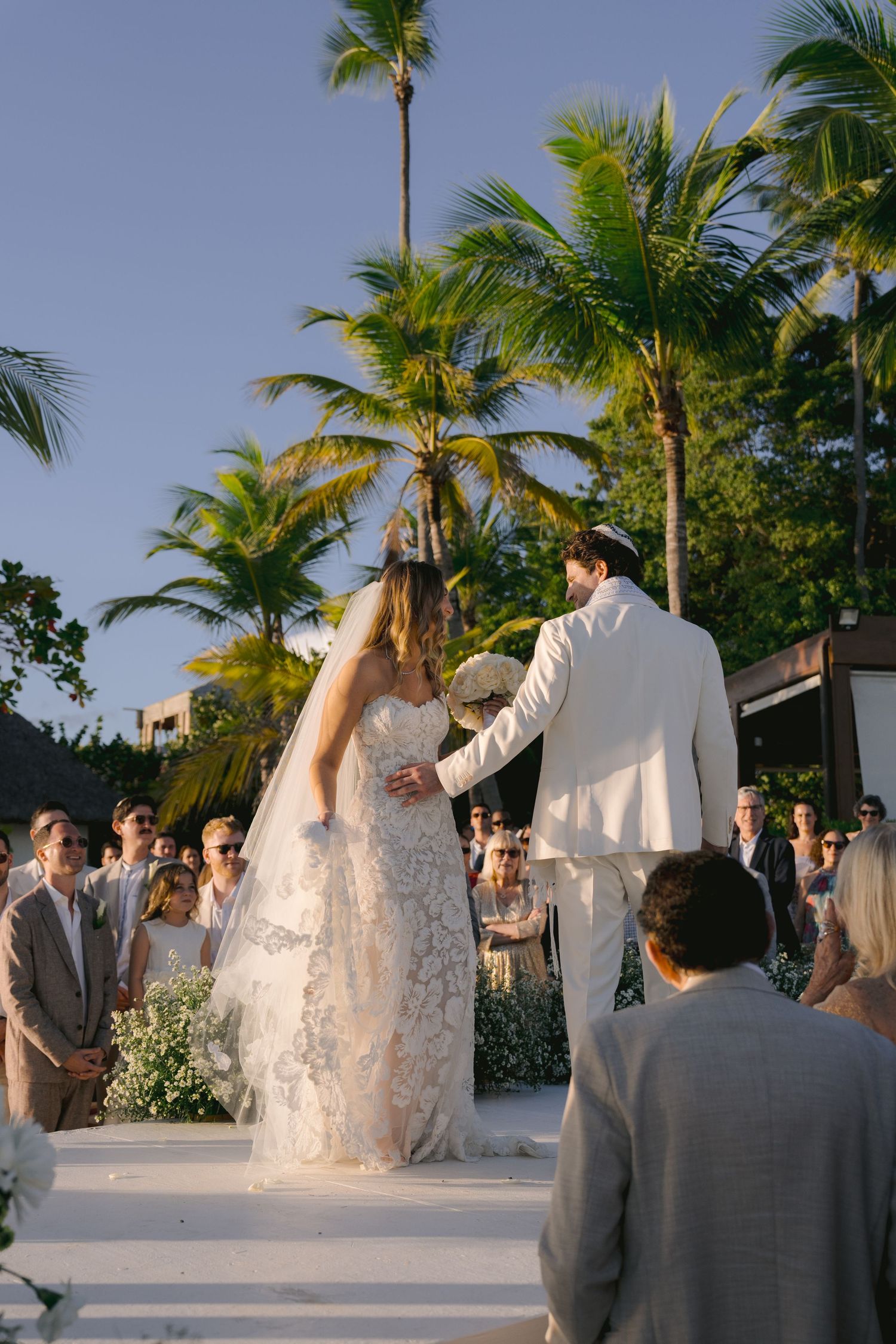 Bride and groom share their first dance surrounded by palm trees at a tropical beach wedding during sunset.