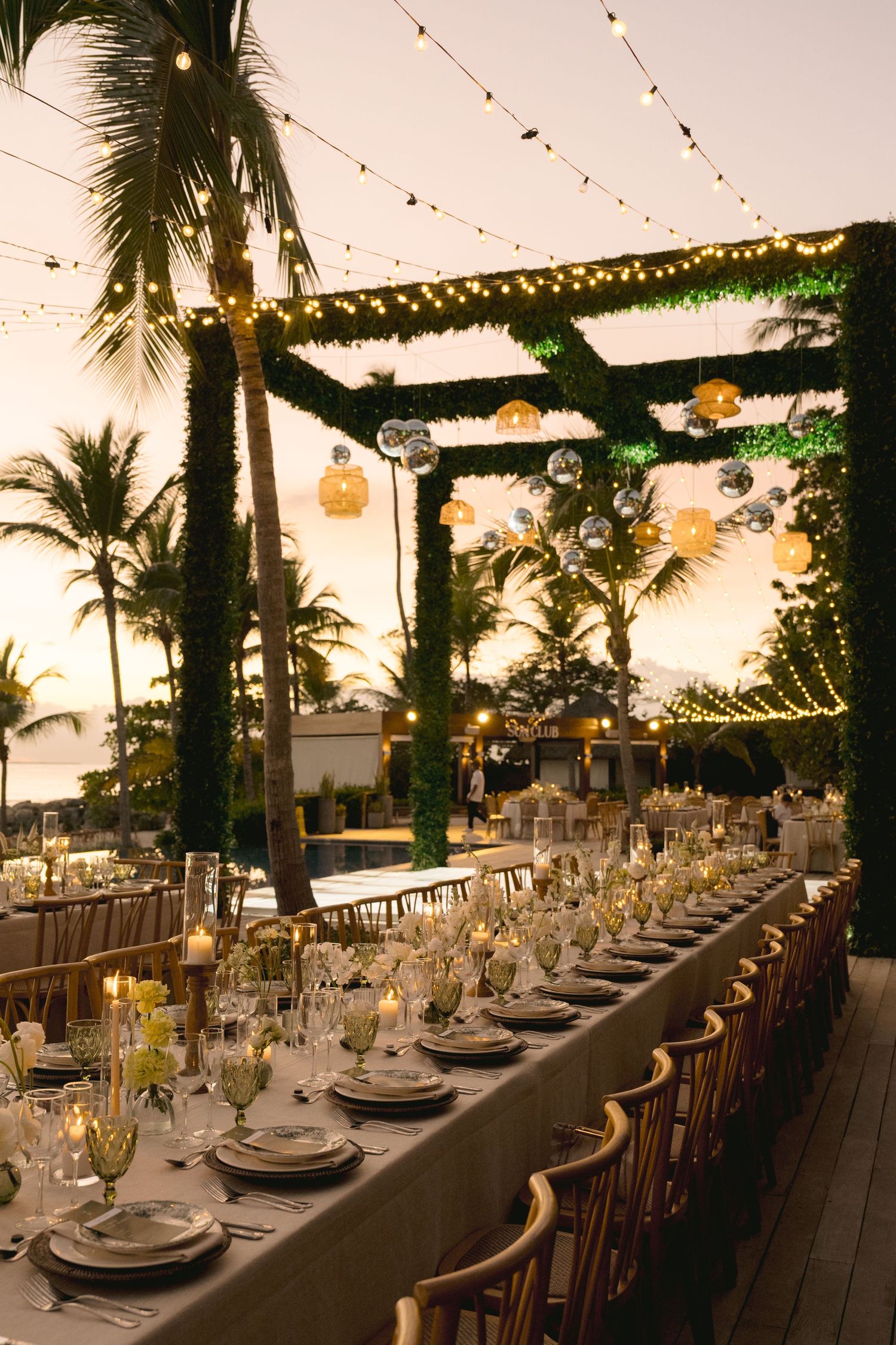 Elegant outdoor wedding reception setup with long tables, string lights and palm trees at sunset creating a tropical ambiance.
