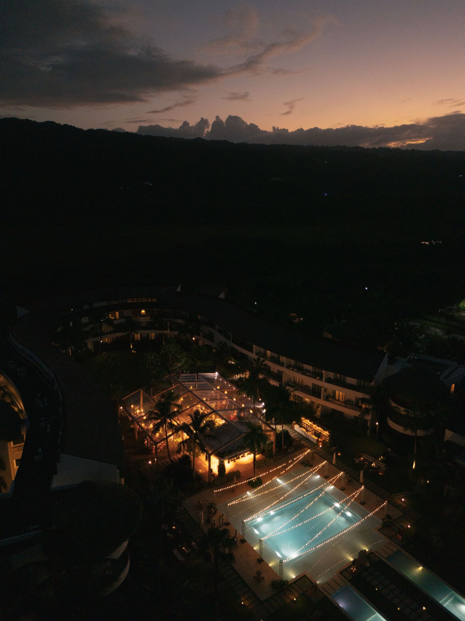 Night view of an illuminated resort pool and courtyard with mountains silhouetted against a sunset sky.