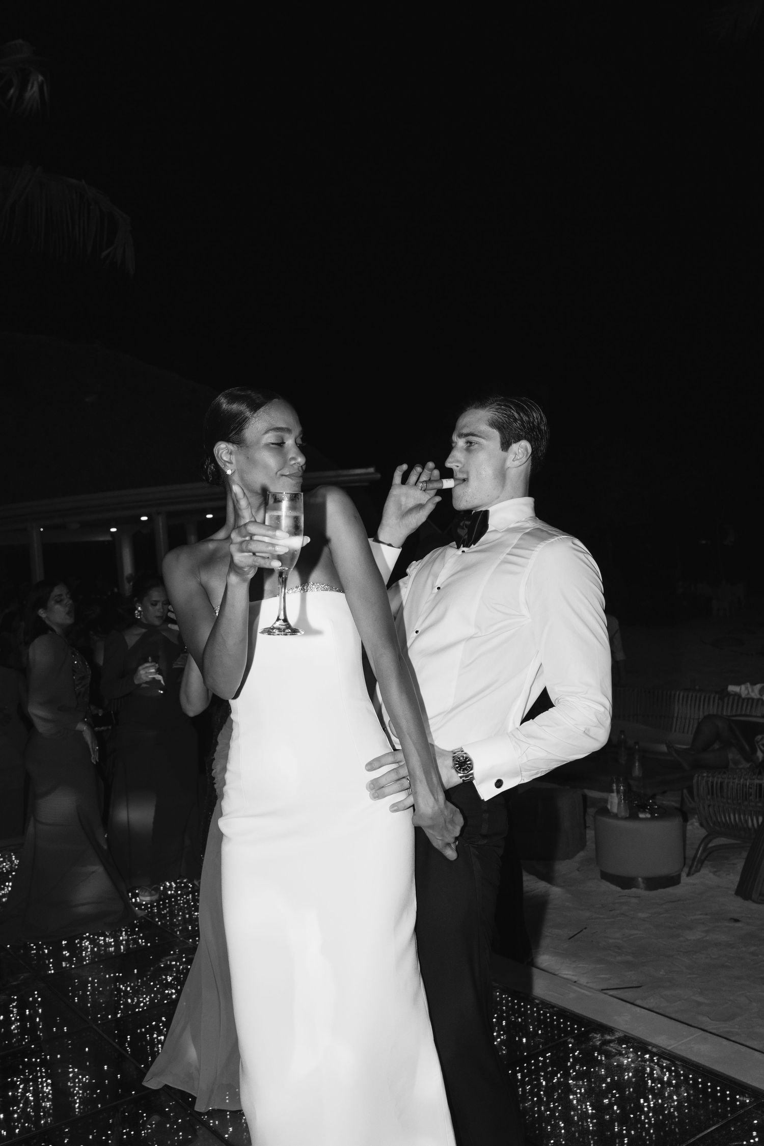 A couple in wedding attire share a drink together at their nighttime reception in black and white photography.