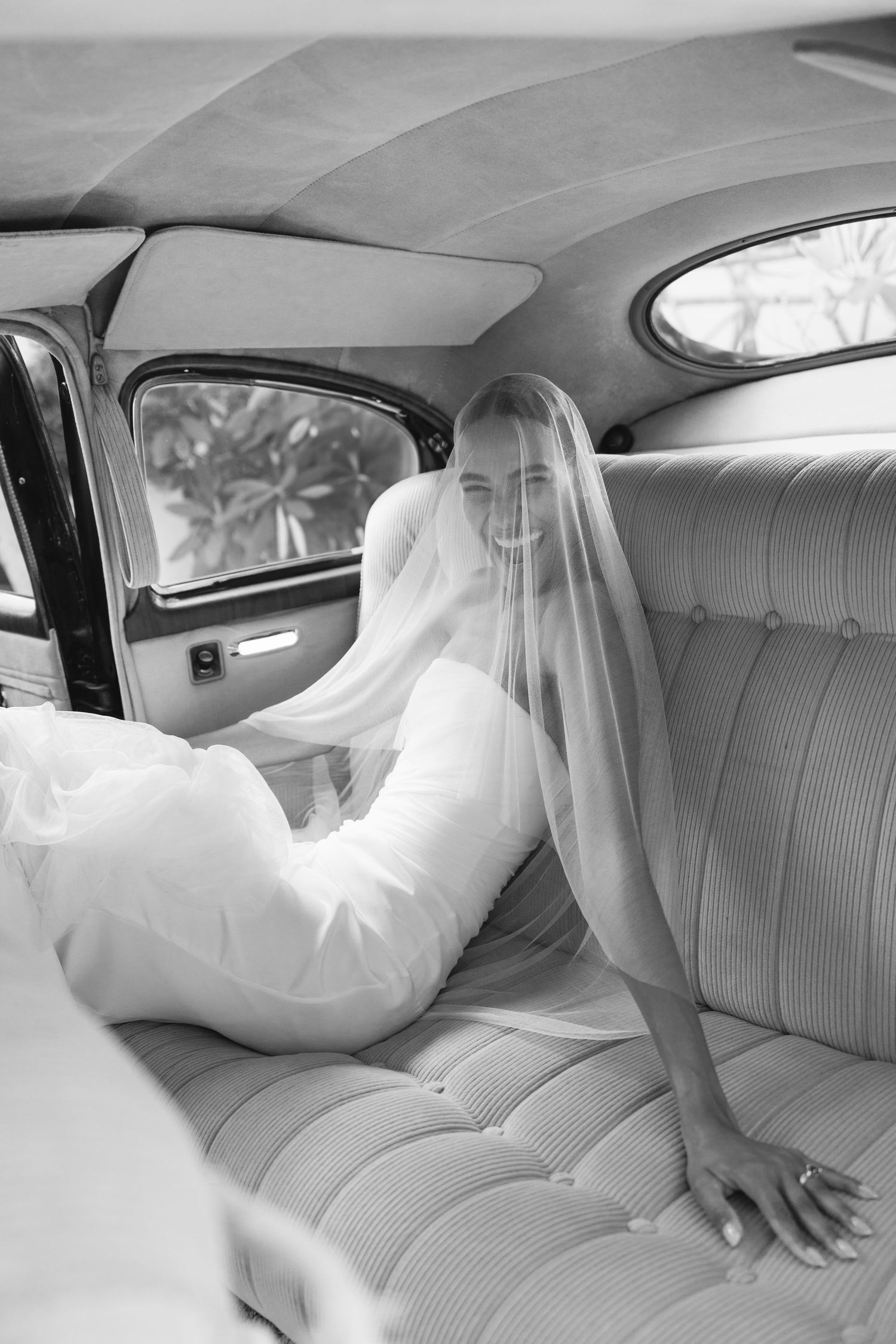 Black and white photo of a bride in a white wedding dress sitting in the backseat of a vintage car.