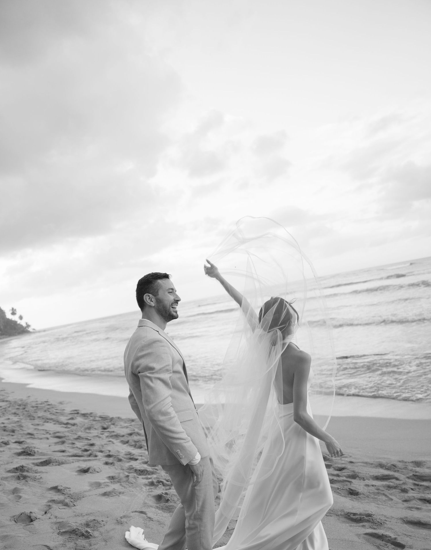 A romantic beach wedding moment captured in black and white with flowing veil on the sand.