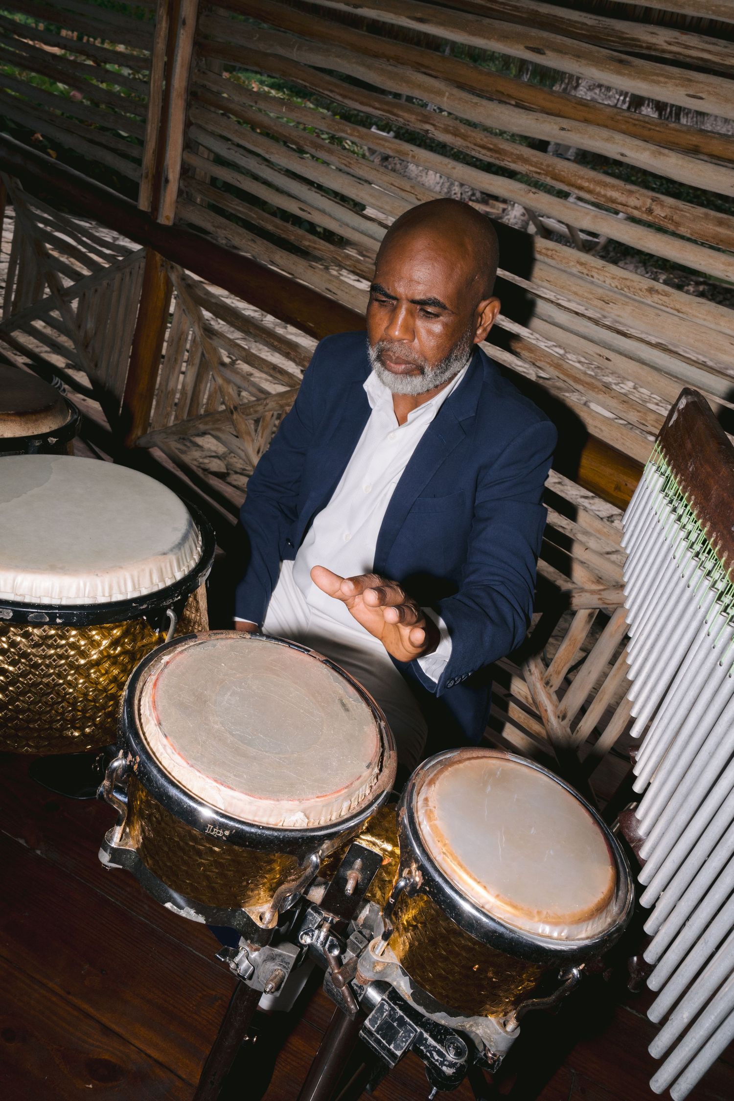 A musician in a navy blazer plays bongo drums in a rustic wooden venue interior.