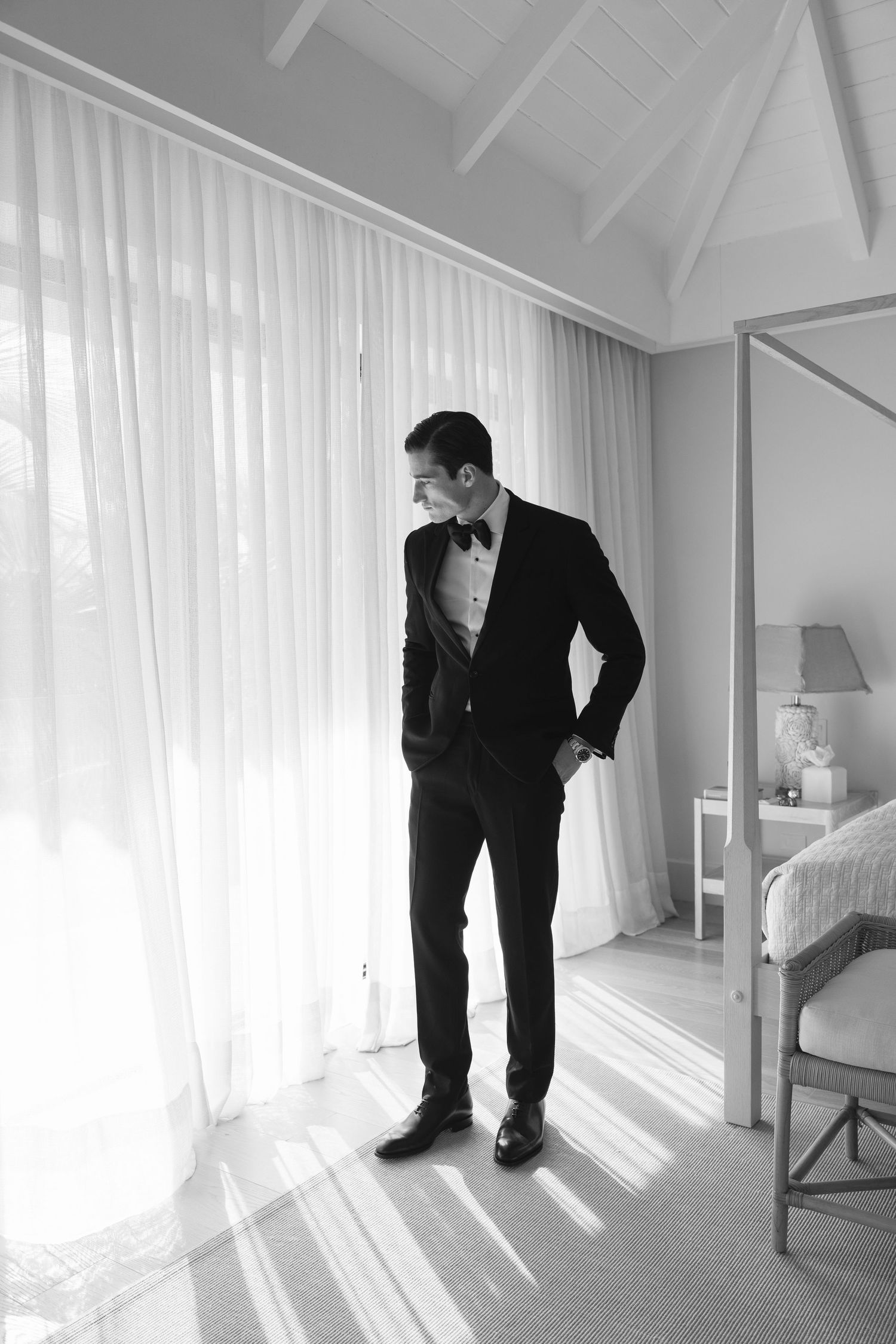 Black and white photograph showing a person in a formal tuxedo and bow tie standing by sheer curtains in a bright room.