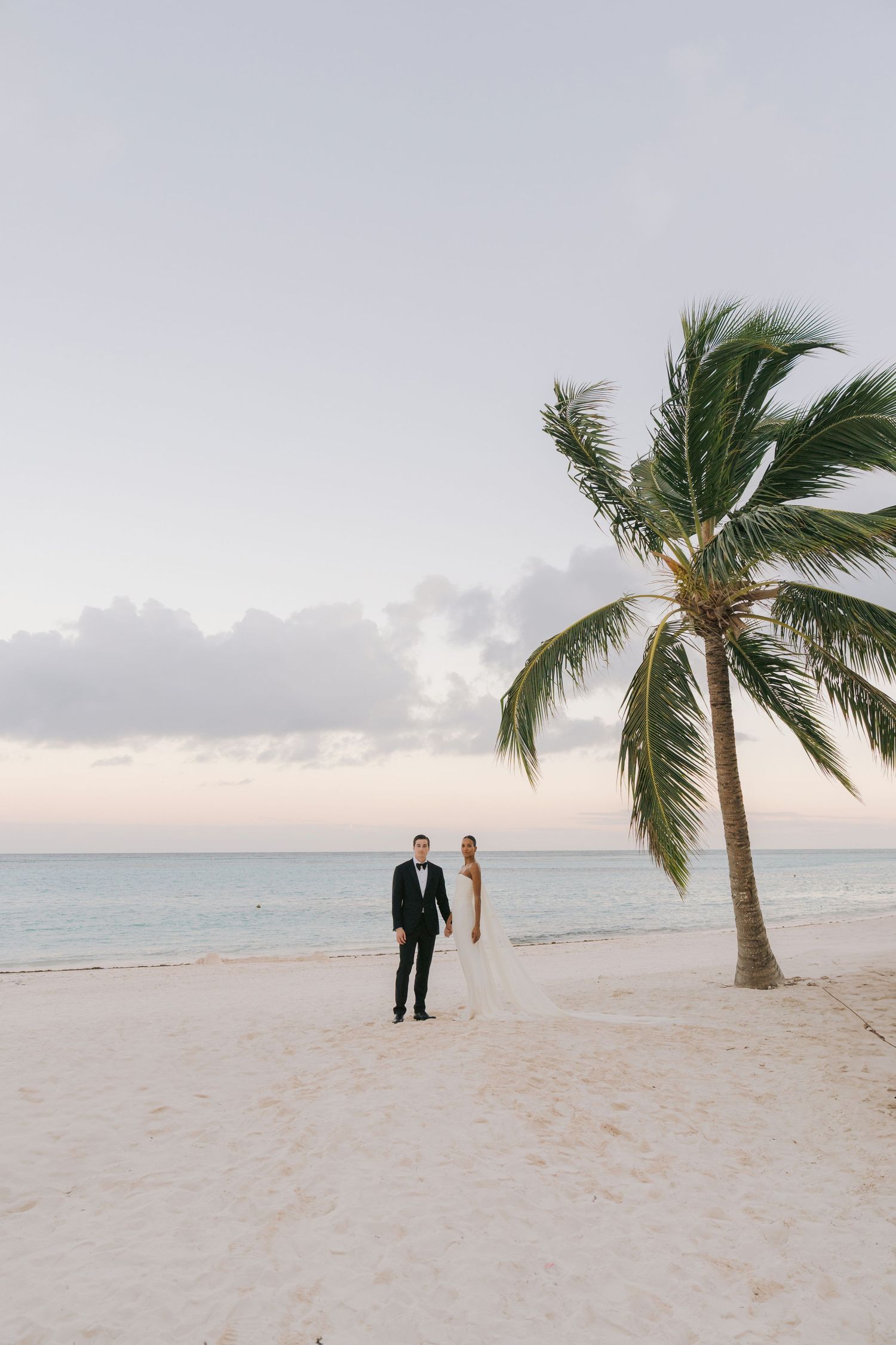 Silhouetted couple stands on pristine white sand beach at sunset with tall palm tree against pastel sky.