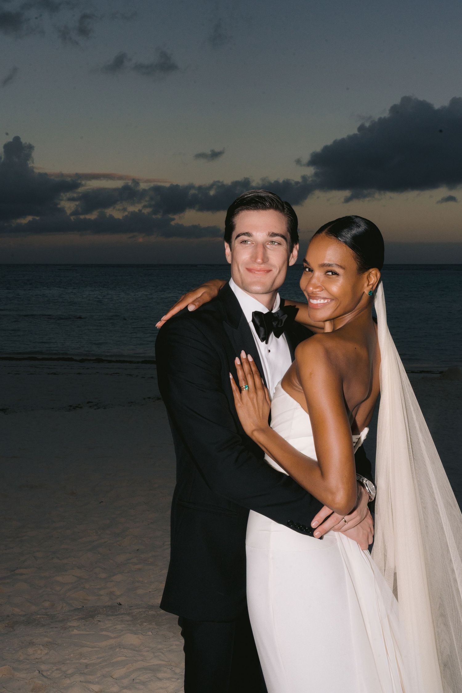 Newlywed couple poses together on beach at sunset with dramatic cloudy sky in background.