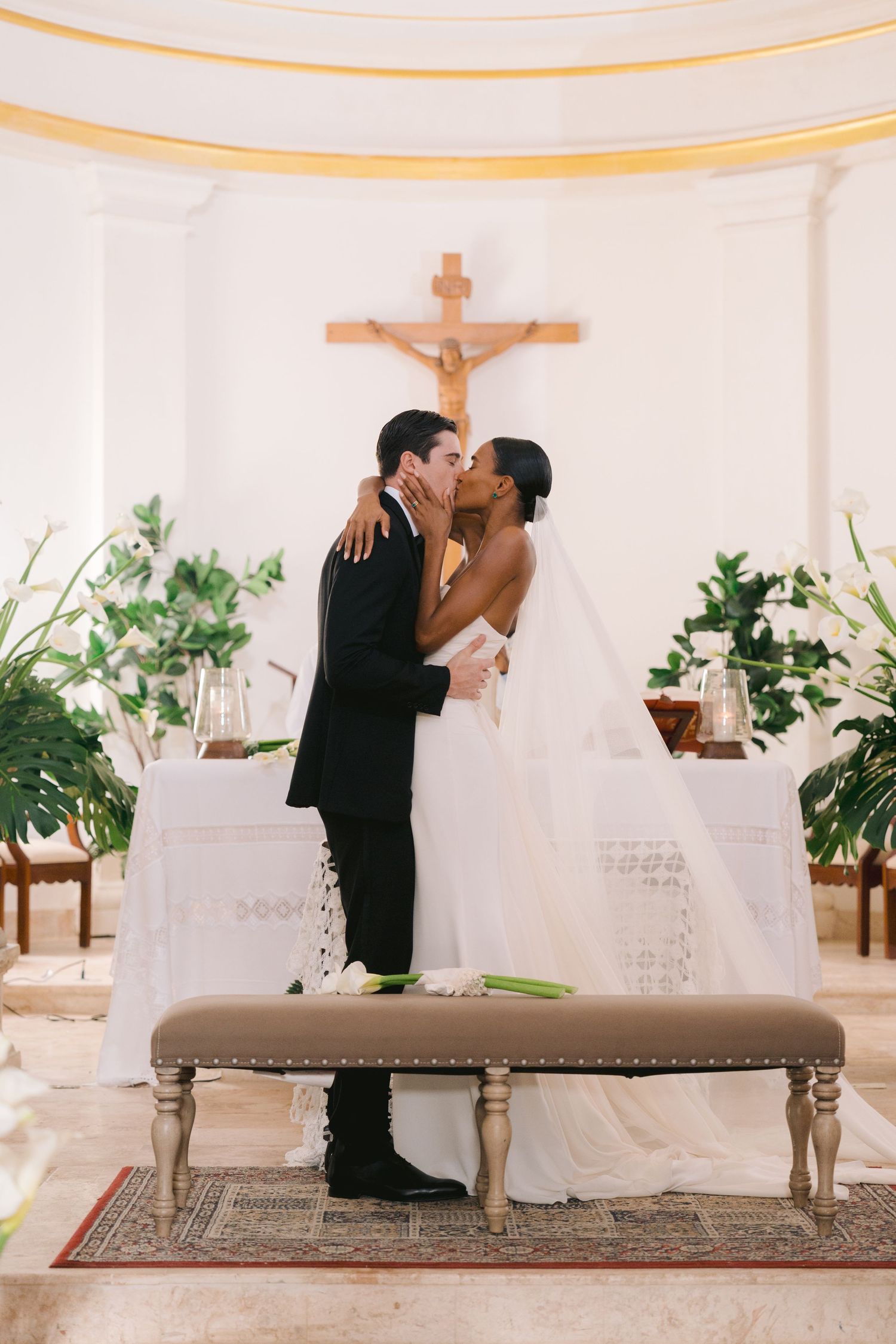 A couple shares their first kiss as newlyweds at the altar of a church decorated with greenery and candles.