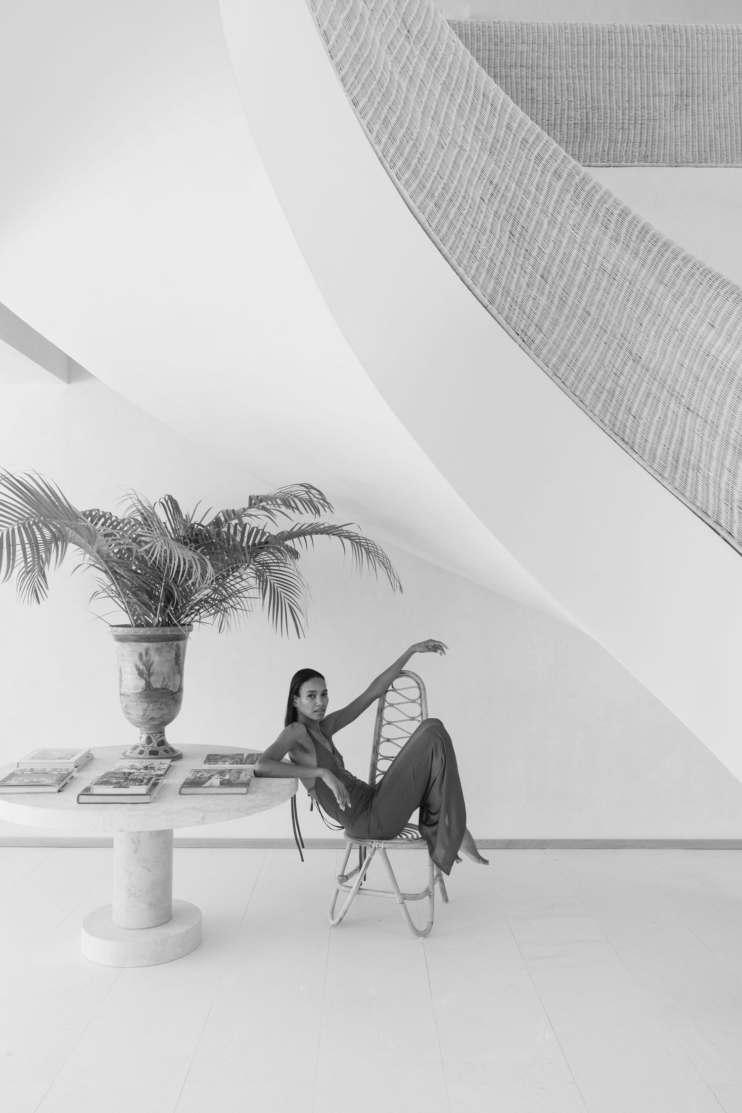 Black and white photograph showing a person lounging in a modern butterfly chair next to a potted palm in a minimalist space.