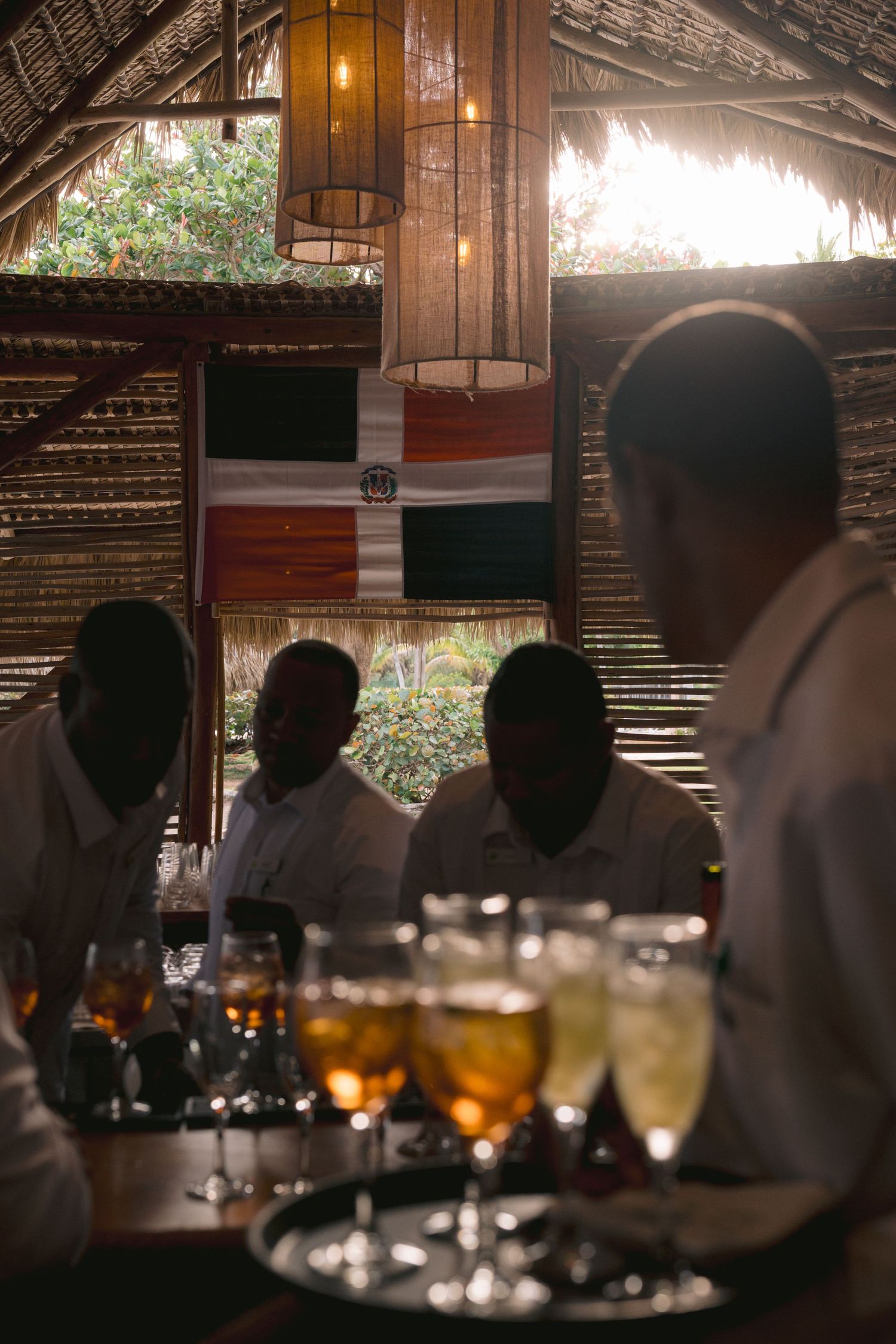 A tiki bar setting with silhouetted figures and wine glasses in the foreground, illuminated by warm lighting.