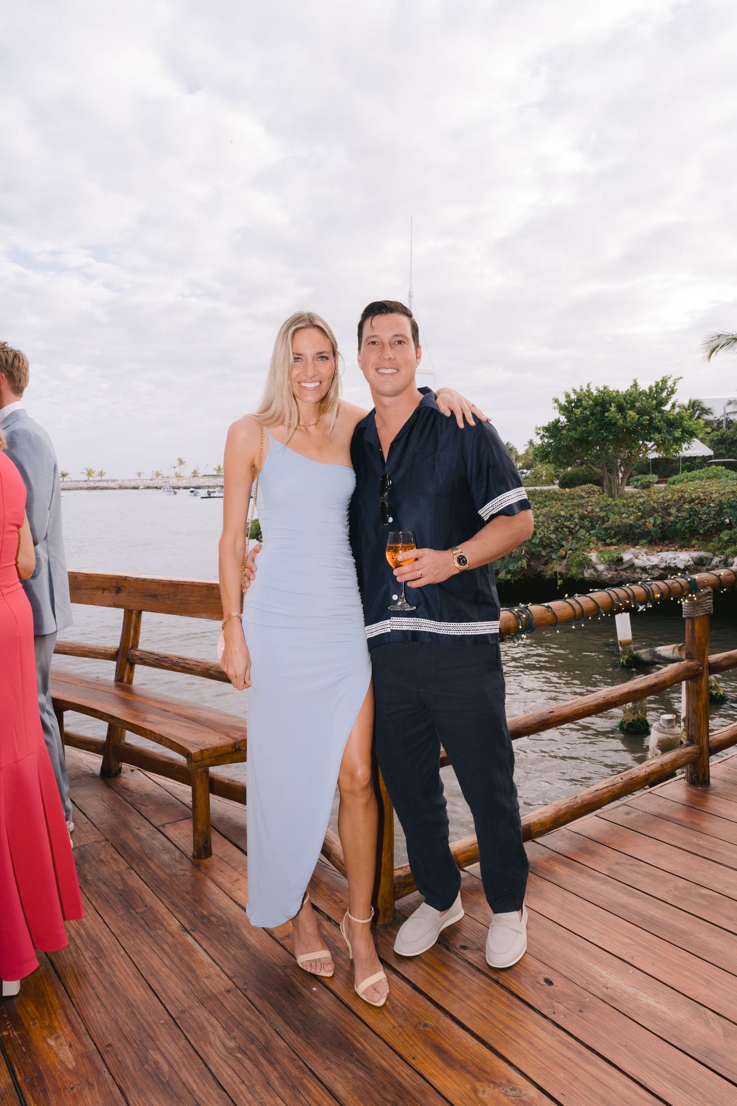 Couple standing together on wooden deck overlooking water, woman wearing light blue dress and man in navy outfit.
