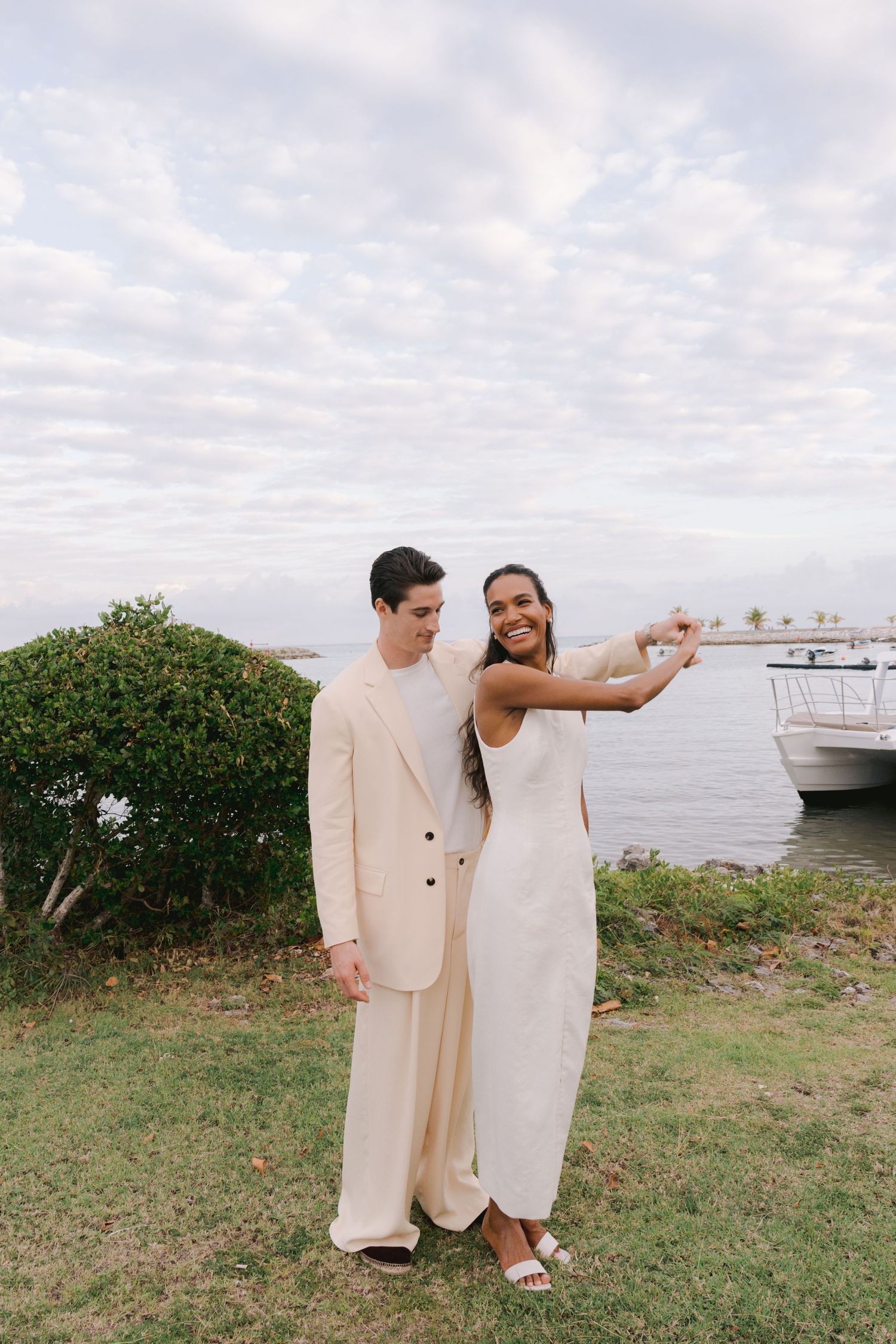 A couple in cream-colored formal attire poses together on a grassy waterfront area with boats visible in the background.