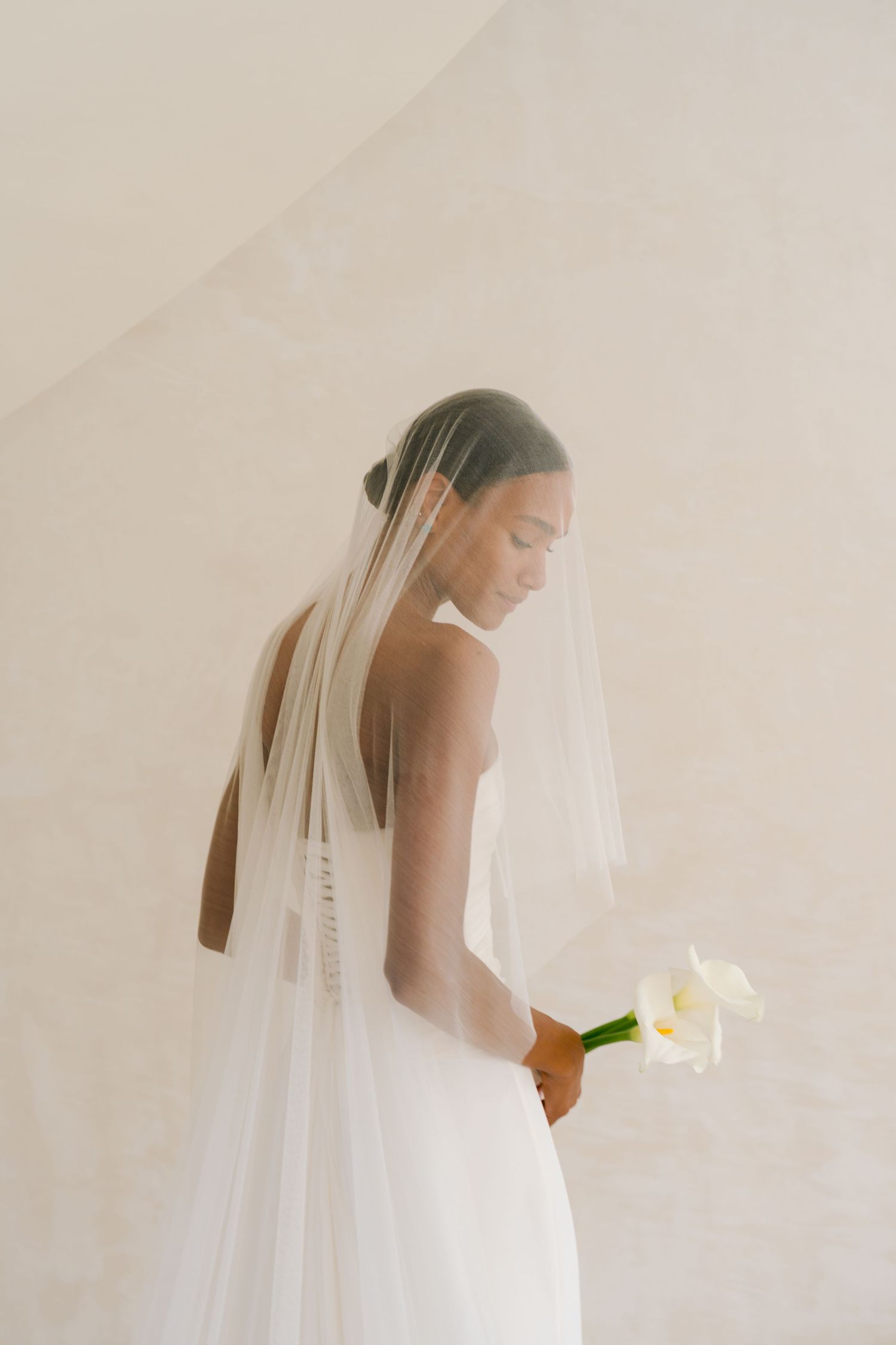 A bride in a flowing white veil and wedding dress holds a small bouquet of white flowers in an ethereal, minimalist setting.