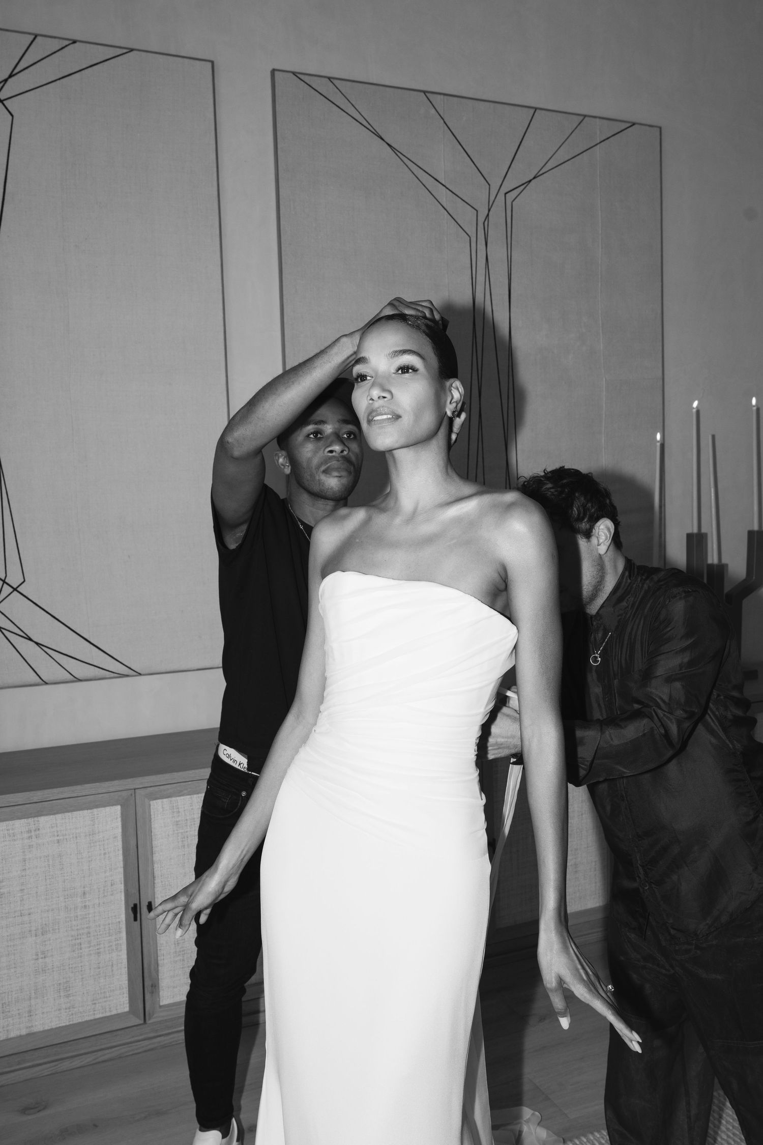 Elegant figure in a white strapless column gown poses gracefully against geometric wall decor at an evening event.