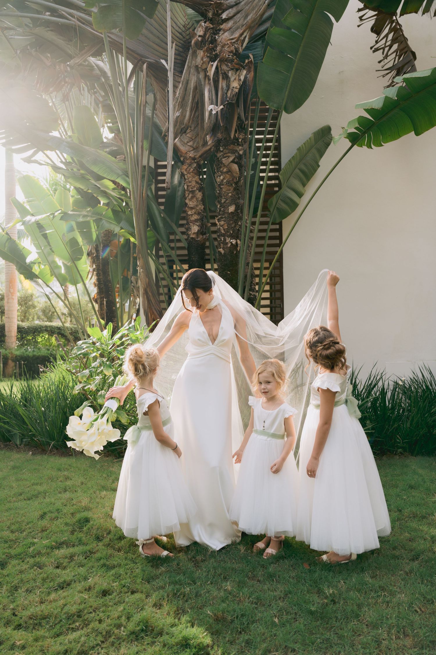 A bride in white gown stands with flower girls in a lush tropical garden setting.