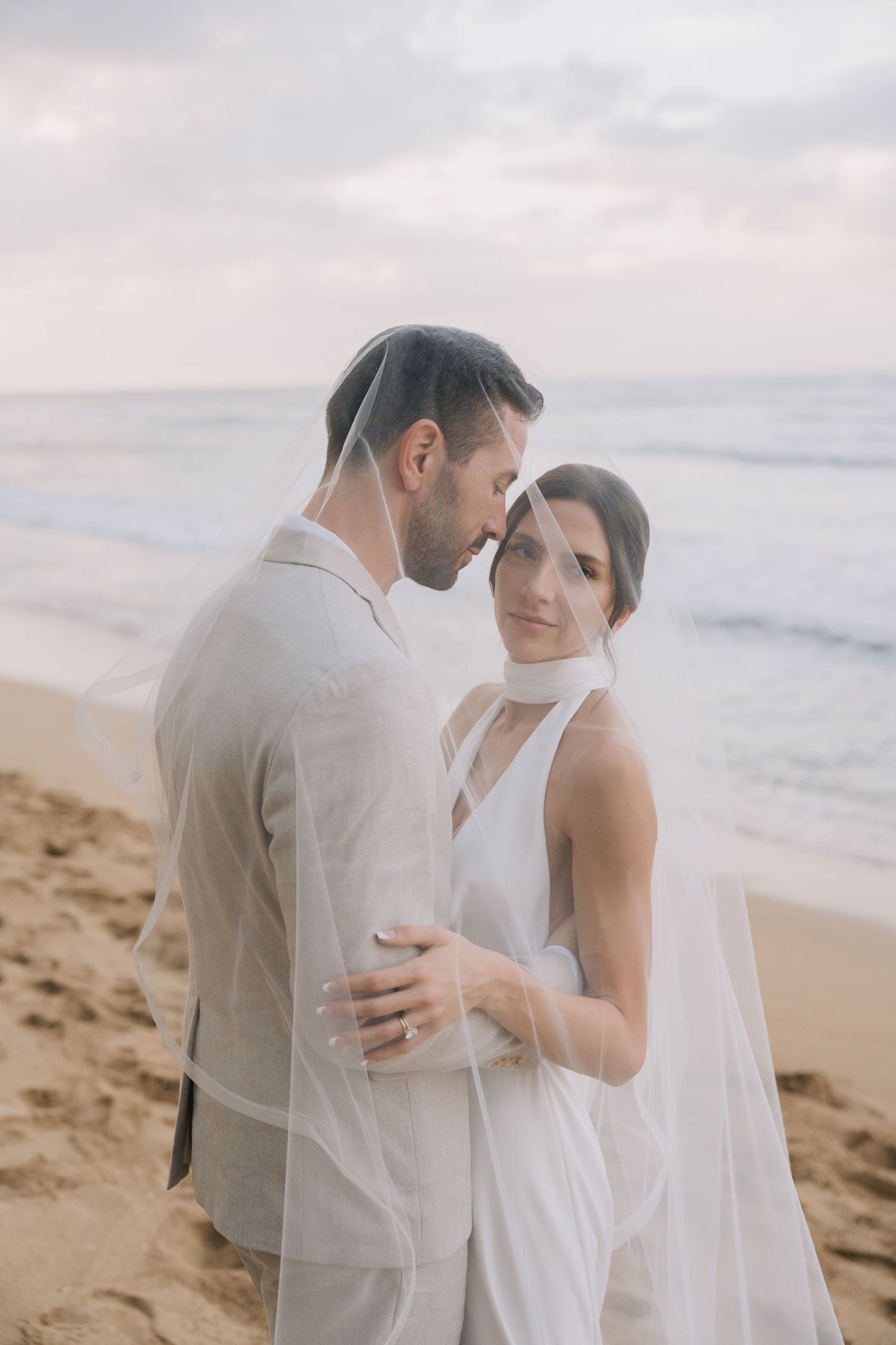 Romantic beach wedding photo of a couple embracing at sunset with flowing veil and ocean backdrop.