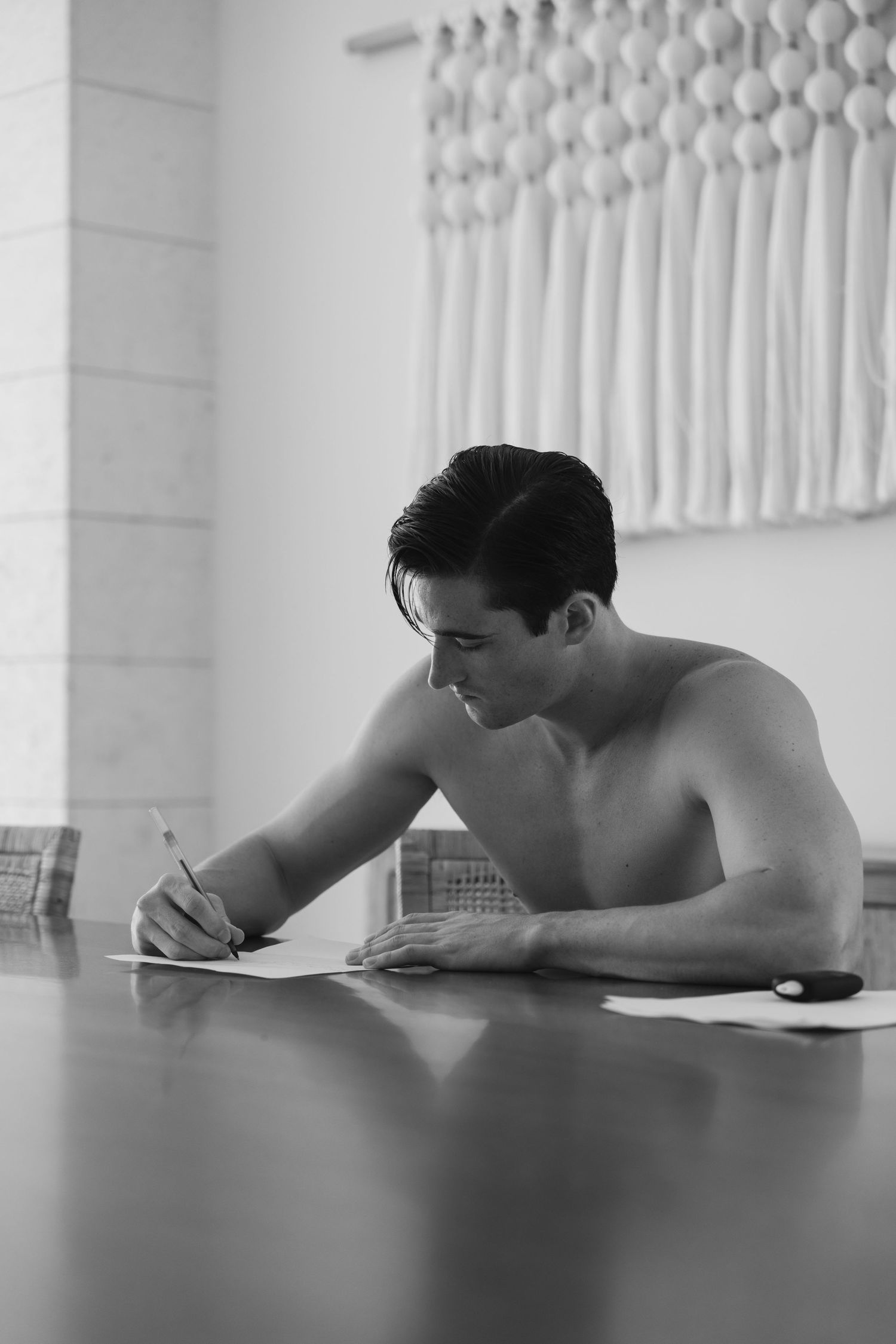 Black and white photo of shirtless figure writing or drawing at table with white sculptural wall decor in background.