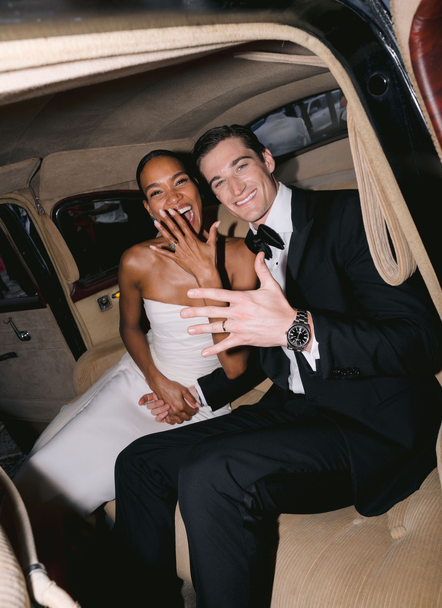 Newlywed couple showing off engagement ring while sitting in back of vintage car after wedding ceremony.