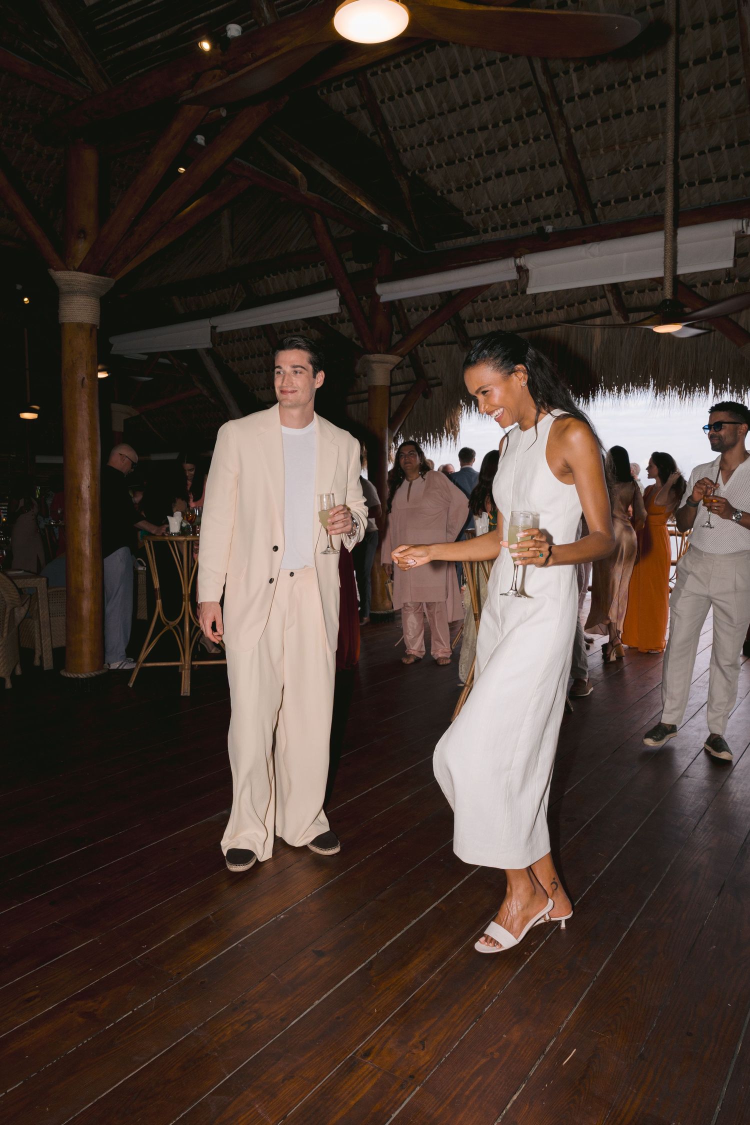 People dancing on a wooden floor at a tropical venue with thatched roof and ambient lighting.