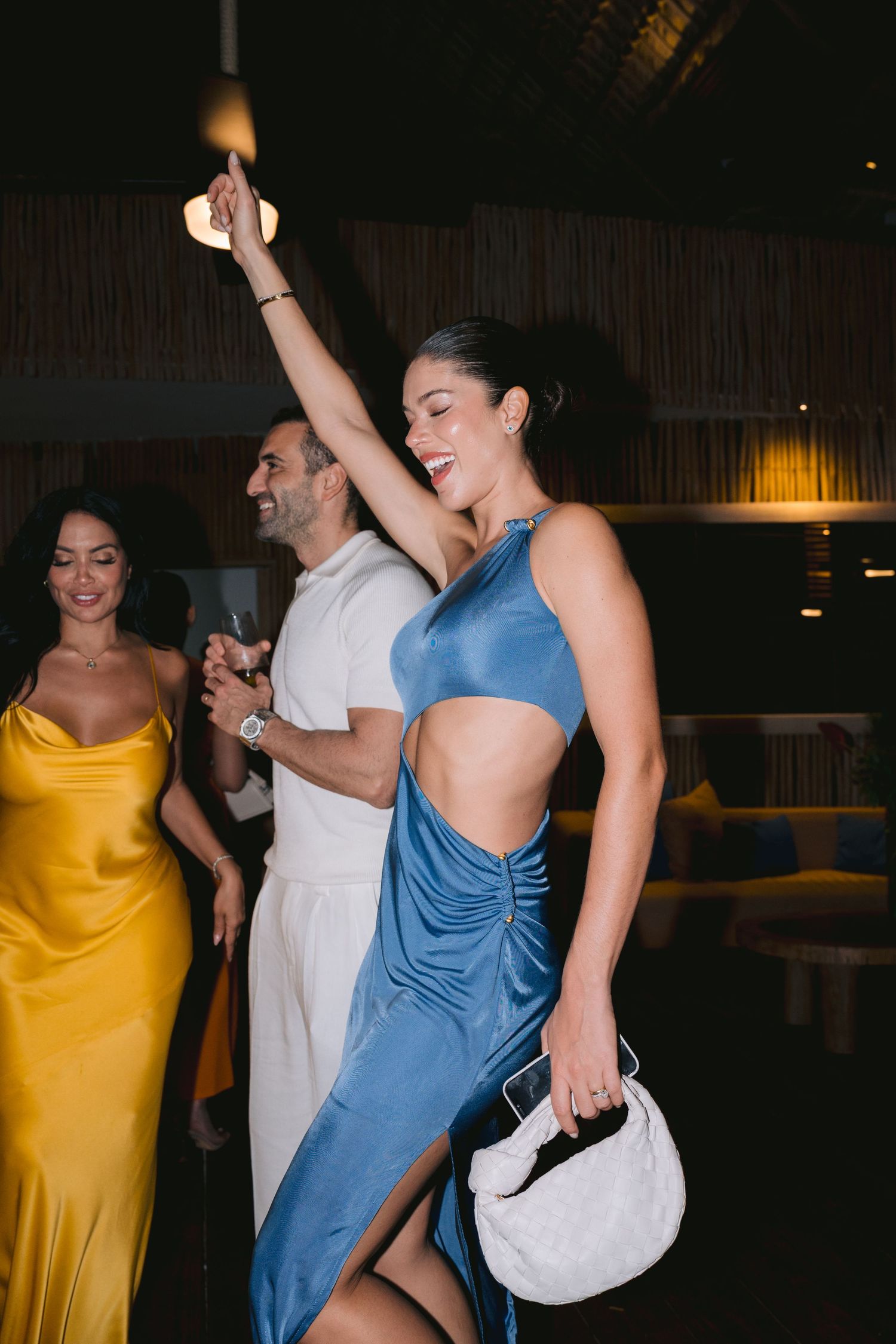 Wedding guests in blue and yellow dresses dance energetically at a dimly lit reception venue.