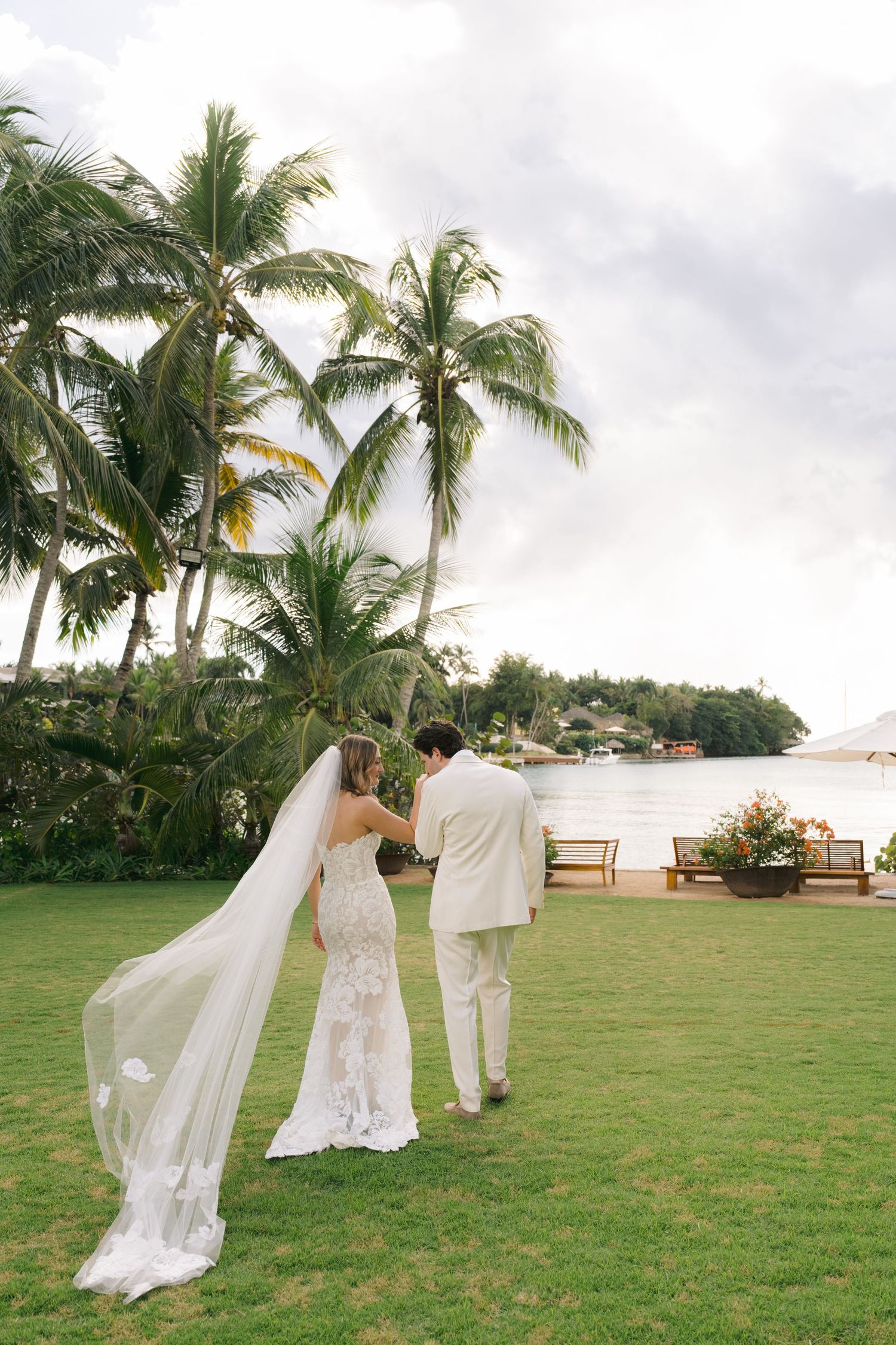 A couple walks hand in hand across a lush green lawn with tropical palm trees and cloudy skies in the background.