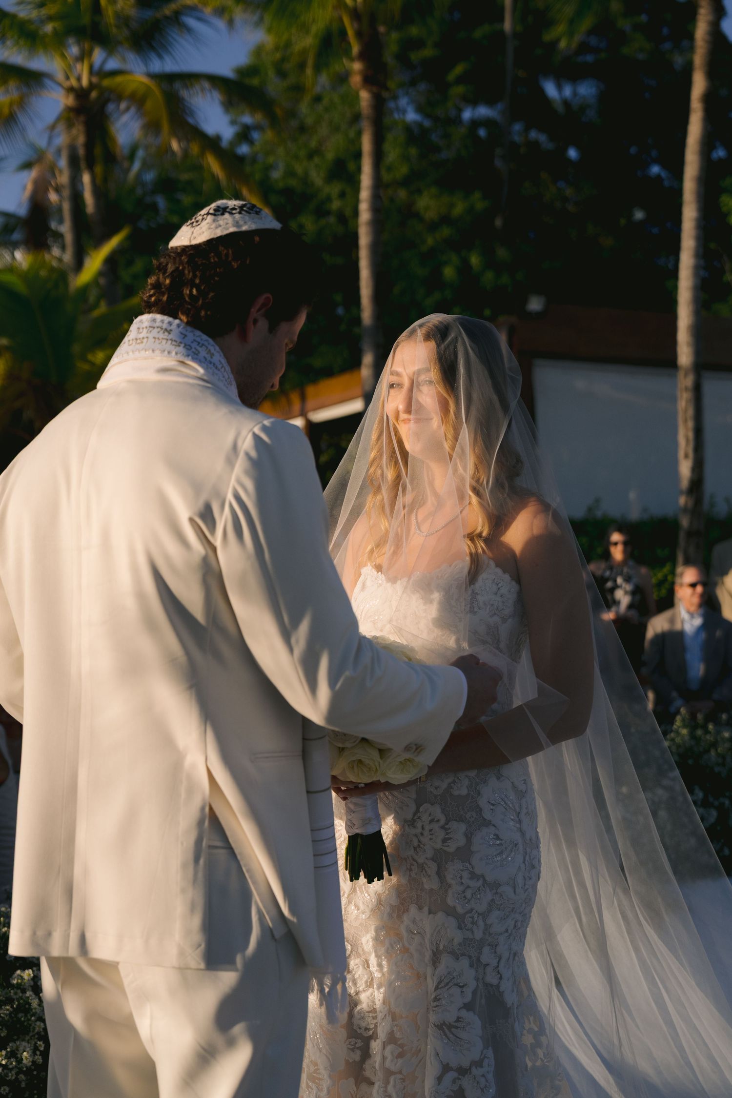 A couple in wedding attire walks along a palm tree-lined path during a tropical sunset ceremony.
