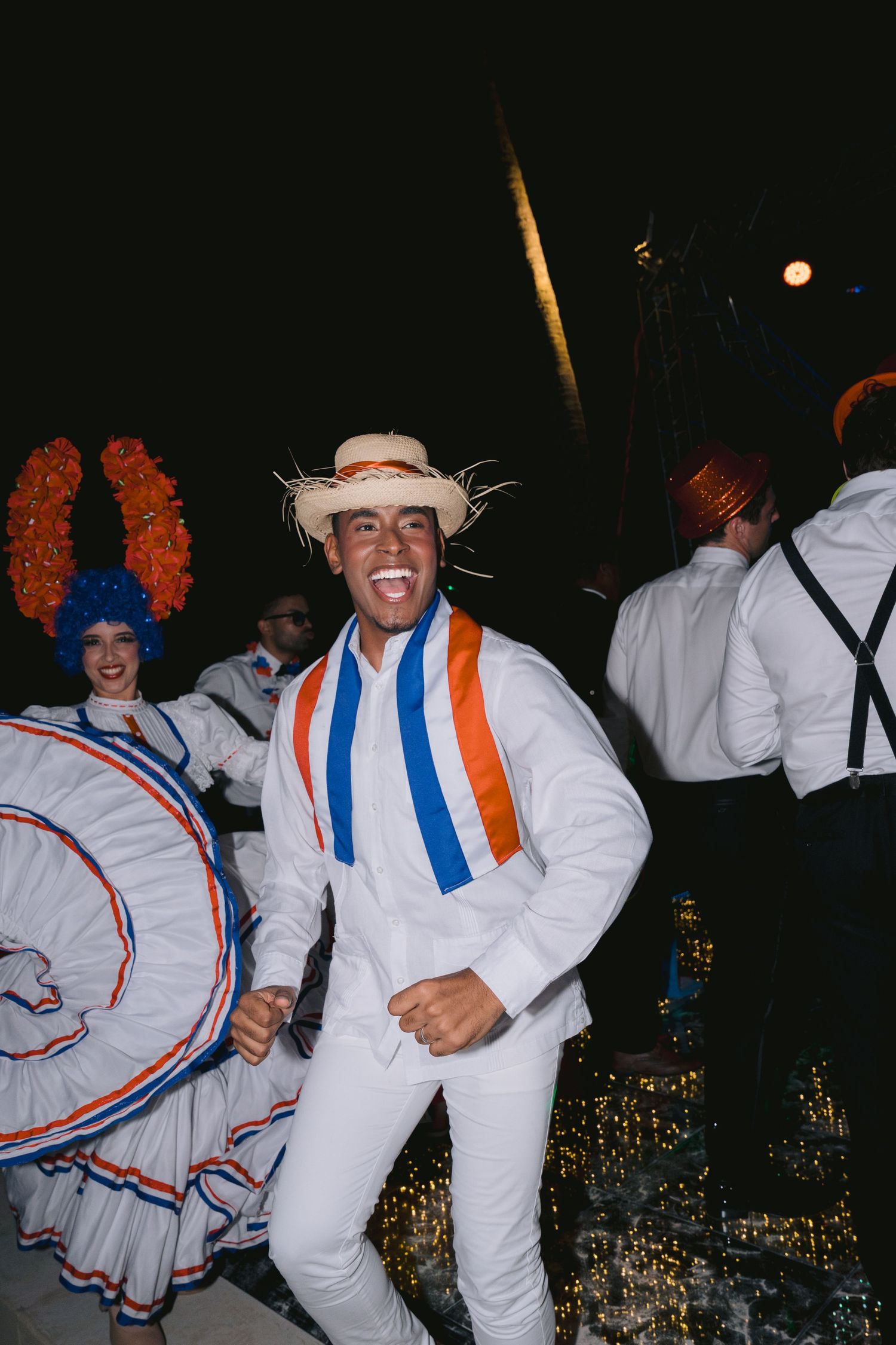 Festively dressed performers posing during a colorful nighttime celebration with white and orange costumes and props.