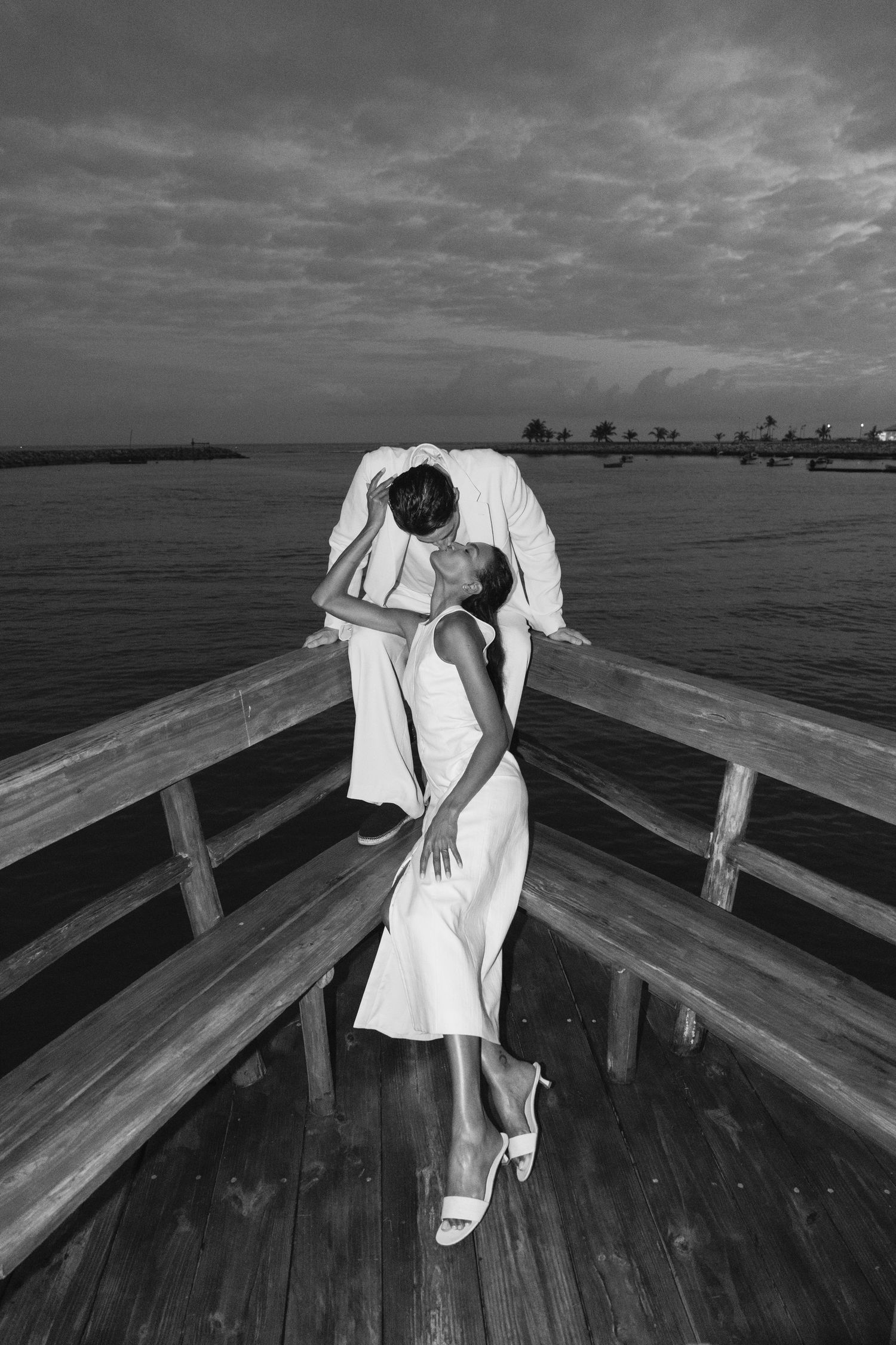 A couple in white clothing embraces romantically on a wooden pier against a dramatic cloudy sky and waterfront view.